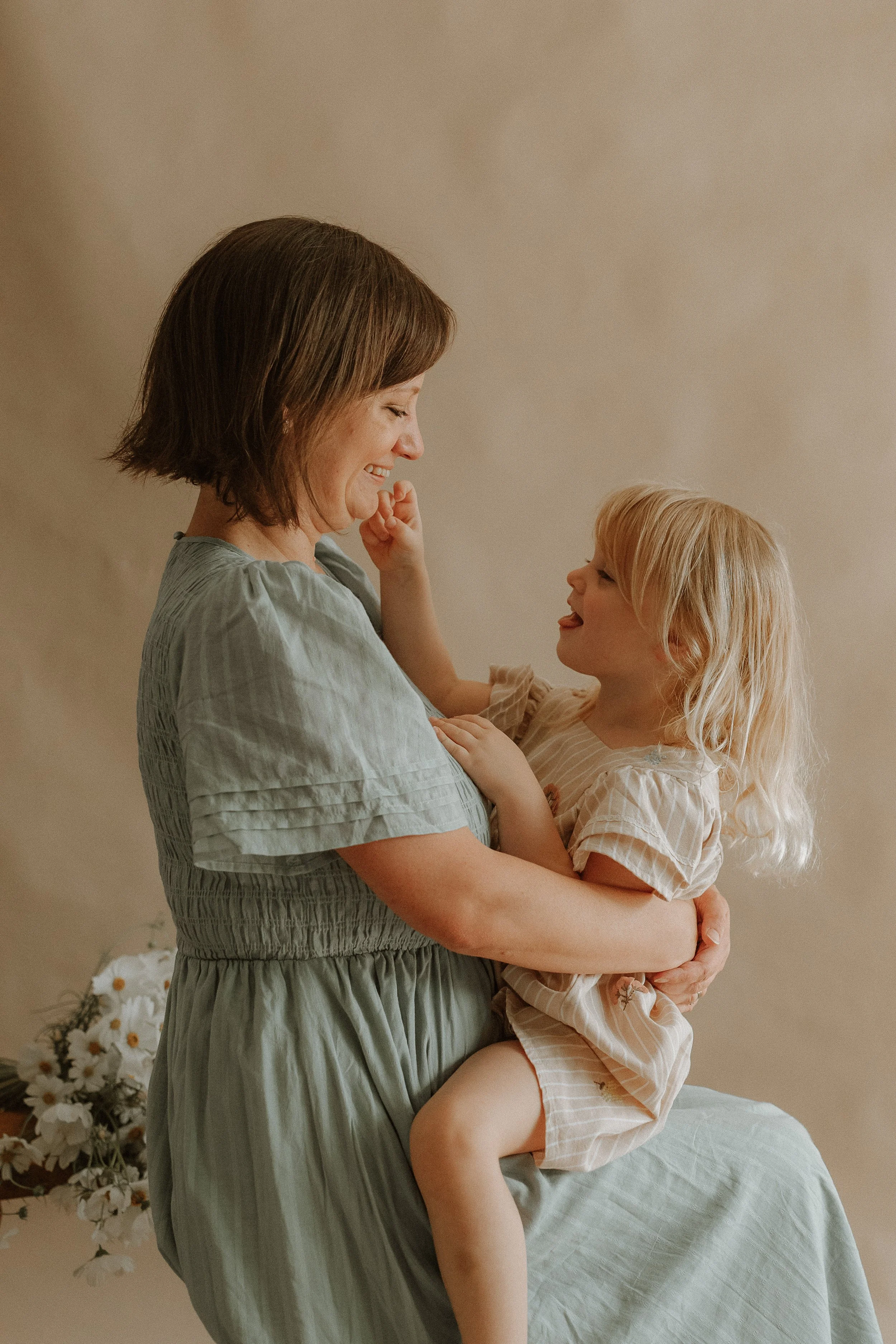 A woman holding a young girl, both smiling and making eye contact, indoors with a neutral wall and white flowers in a vase in the background.