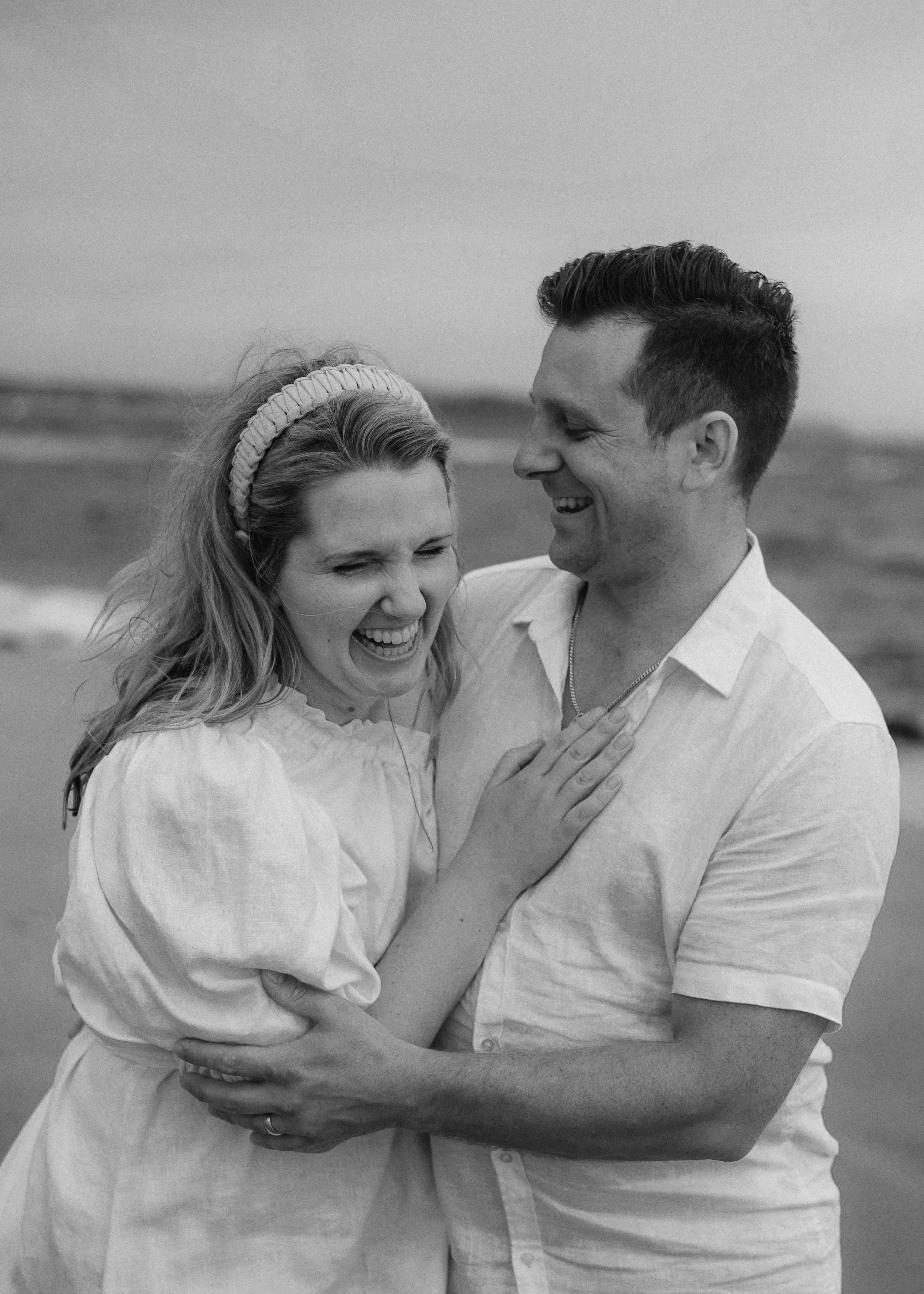A happy couple embracing each other on a beach, both smiling and looking at each other in black and white.