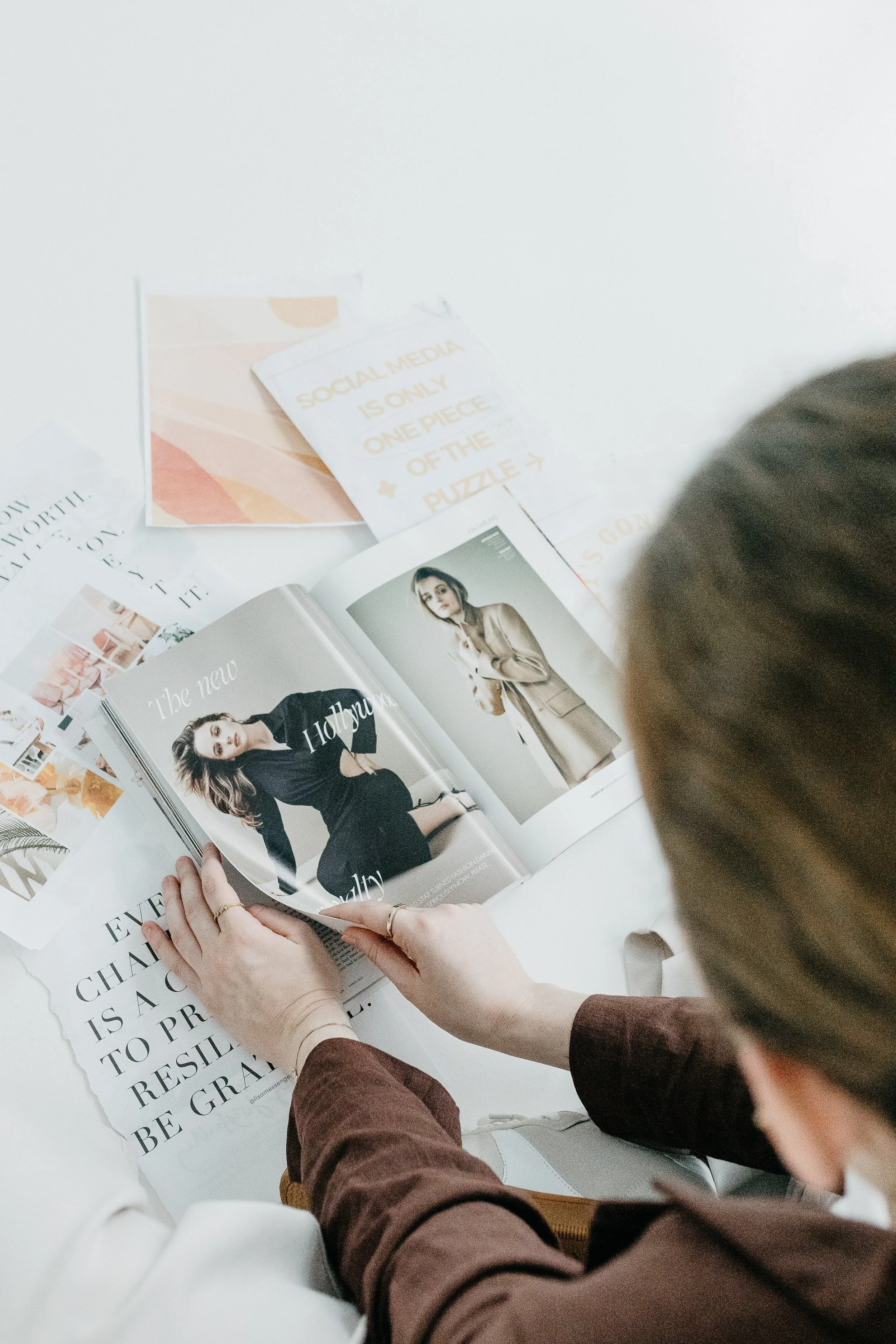A person with long hair, wearing a brown long-sleeve shirt, is flipping through a magazine on a table filled with various printed materials and advertisements.
