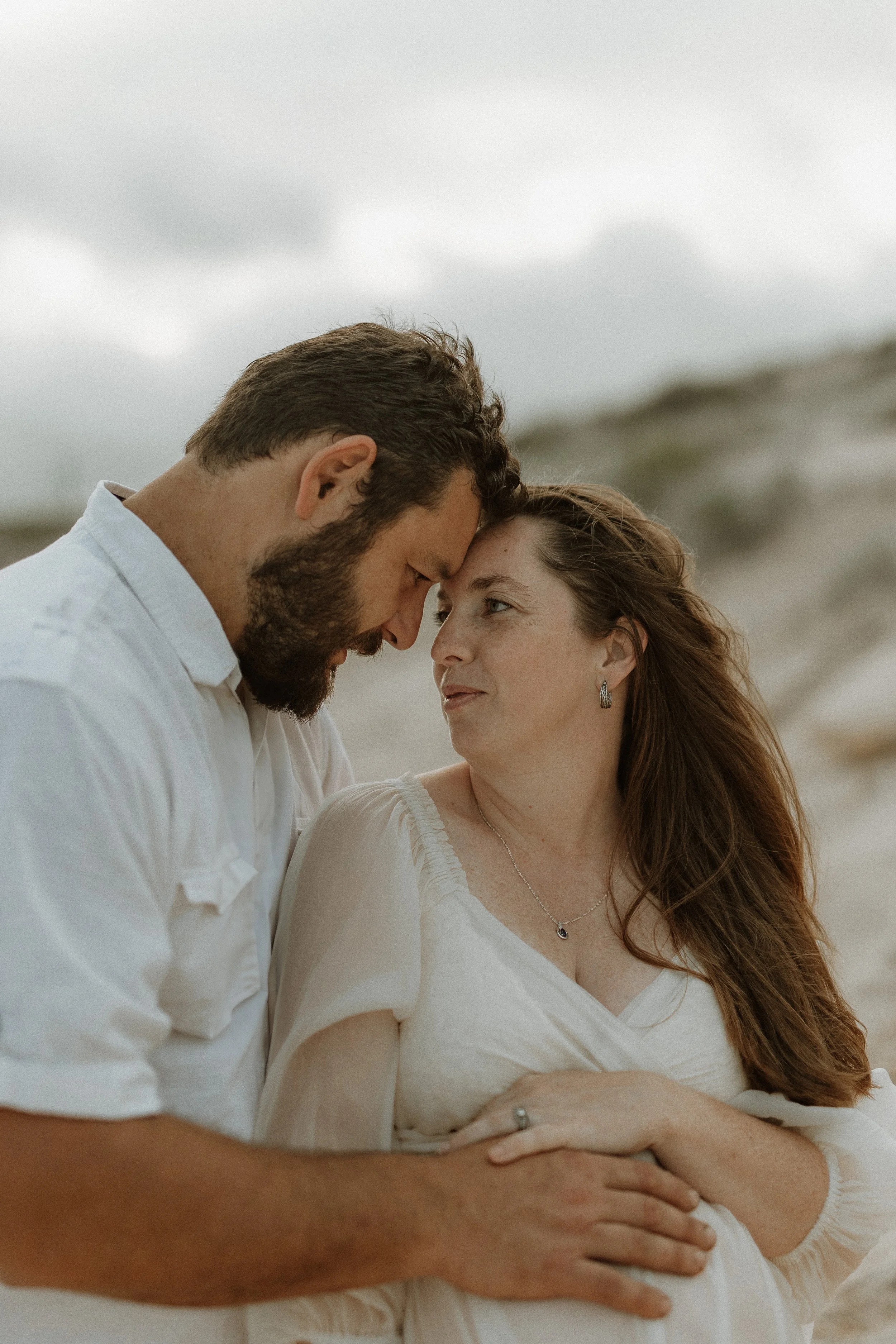 A couple is standing close together on a beach, touching foreheads and looking into each other's eyes, with a cloudy sky in the background.