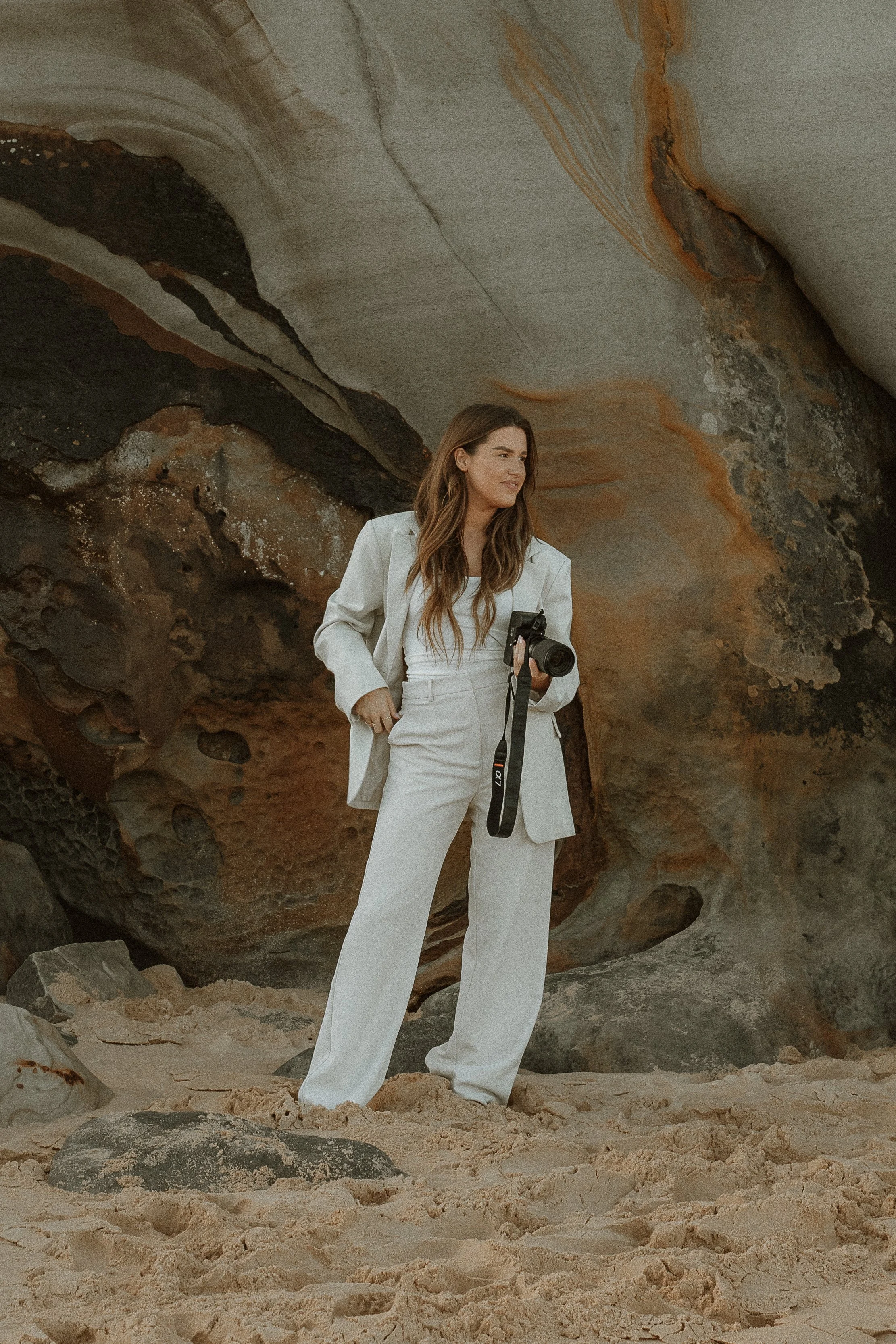 A young woman in white pants and a white blazer holding a camera stands on a sandy beach with large rocks and a rocky cliff behind her.