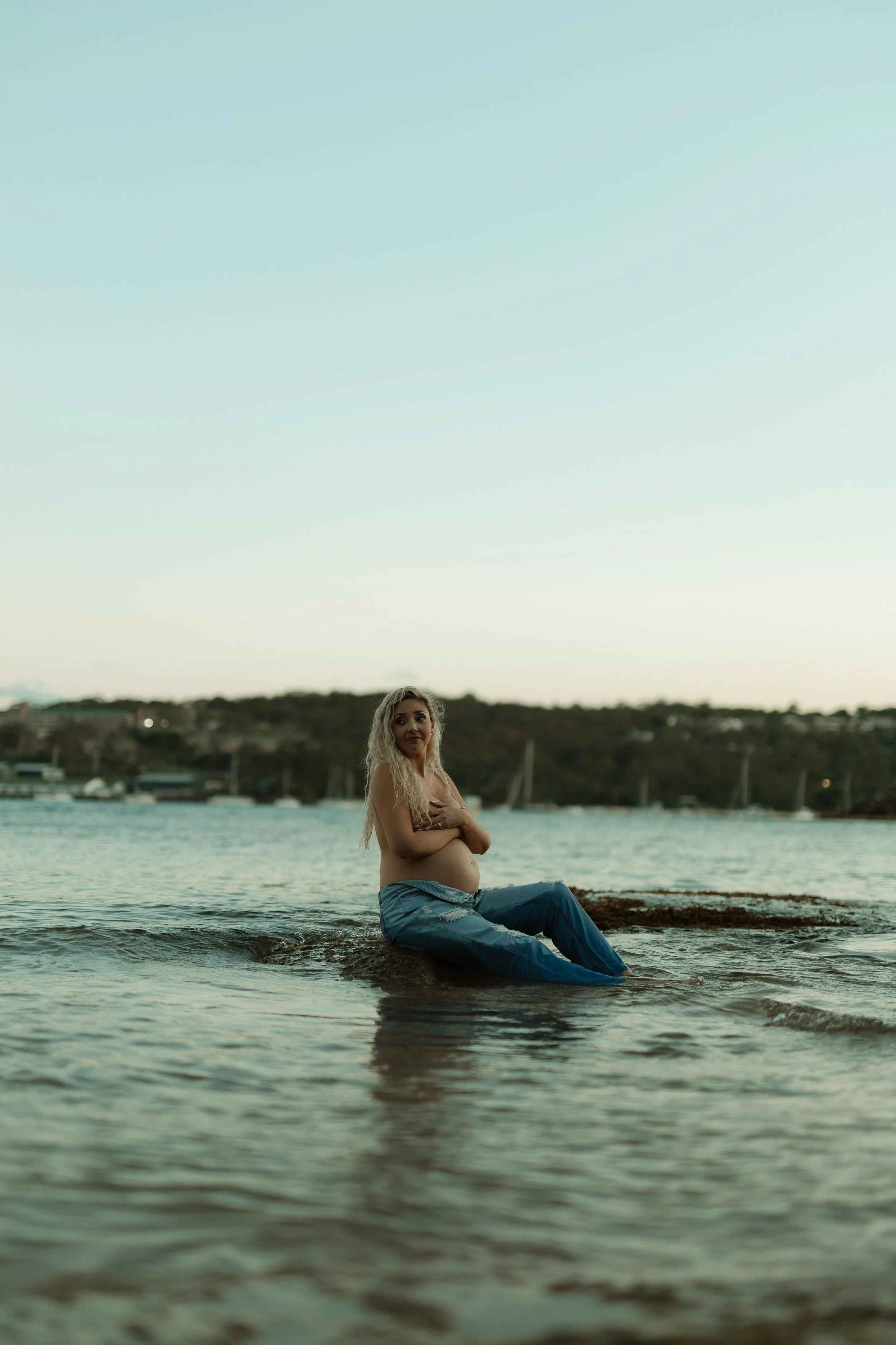 A pregnant woman with long blonde hair sitting in shallow water at a beach during sunset, wearing jeans and embracing her belly, with boats and shoreline in the background.