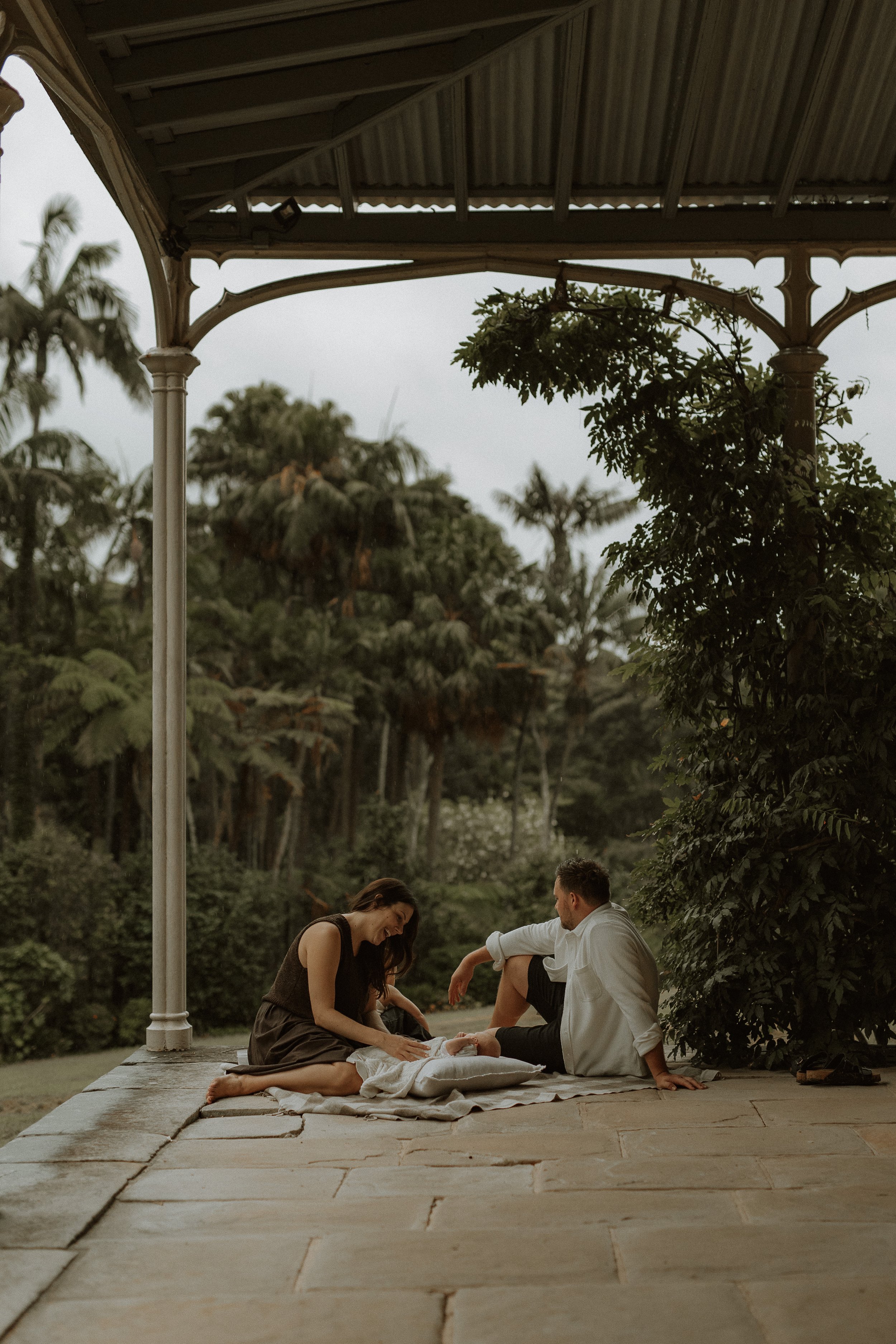 A couple and their children sitting on a blanket outside on a stone patio, having a discussion in a park with trees and greenery around, under a metal roofed structure.