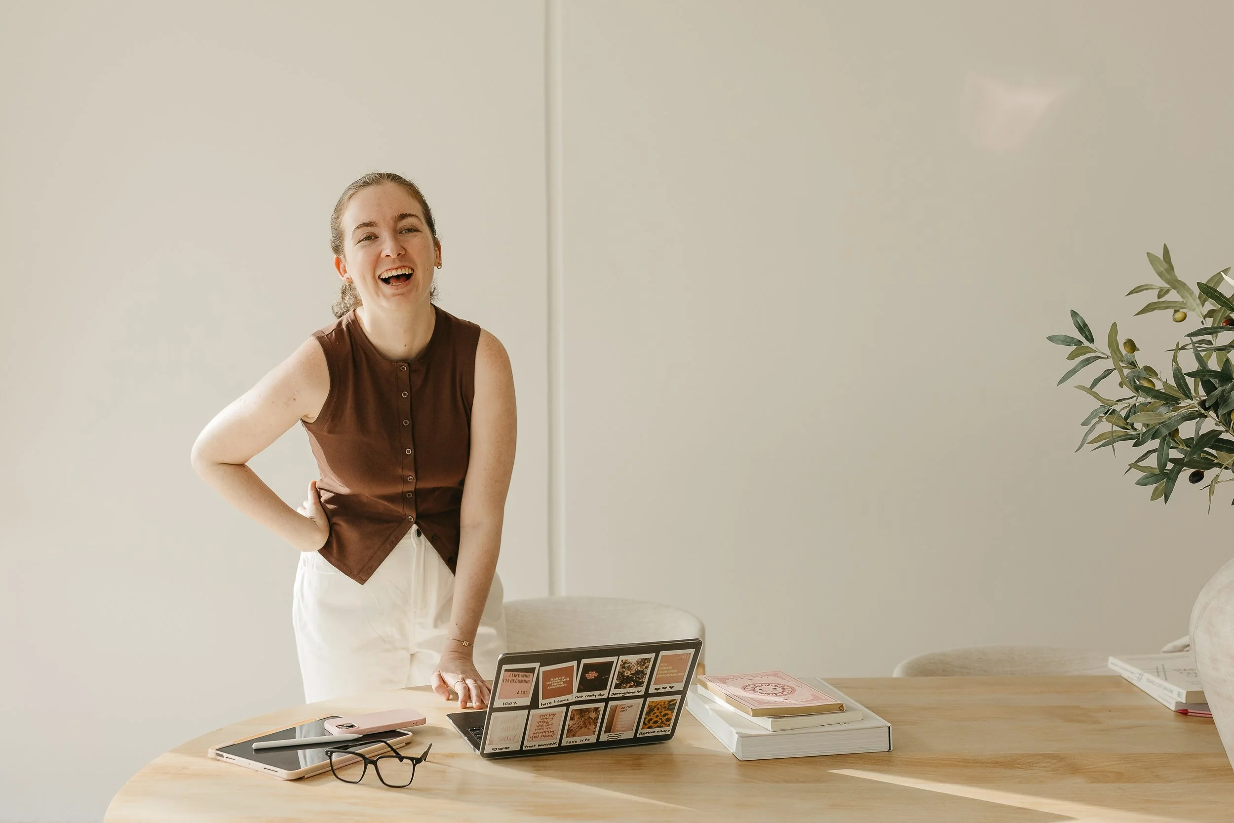 A woman with brown hair tied back, wearing a sleeveless brown top and white pants, is smiling and standing at a table in a minimalist room with a large green plant nearby. The table has various laptops, books, papers, and glasses on it.