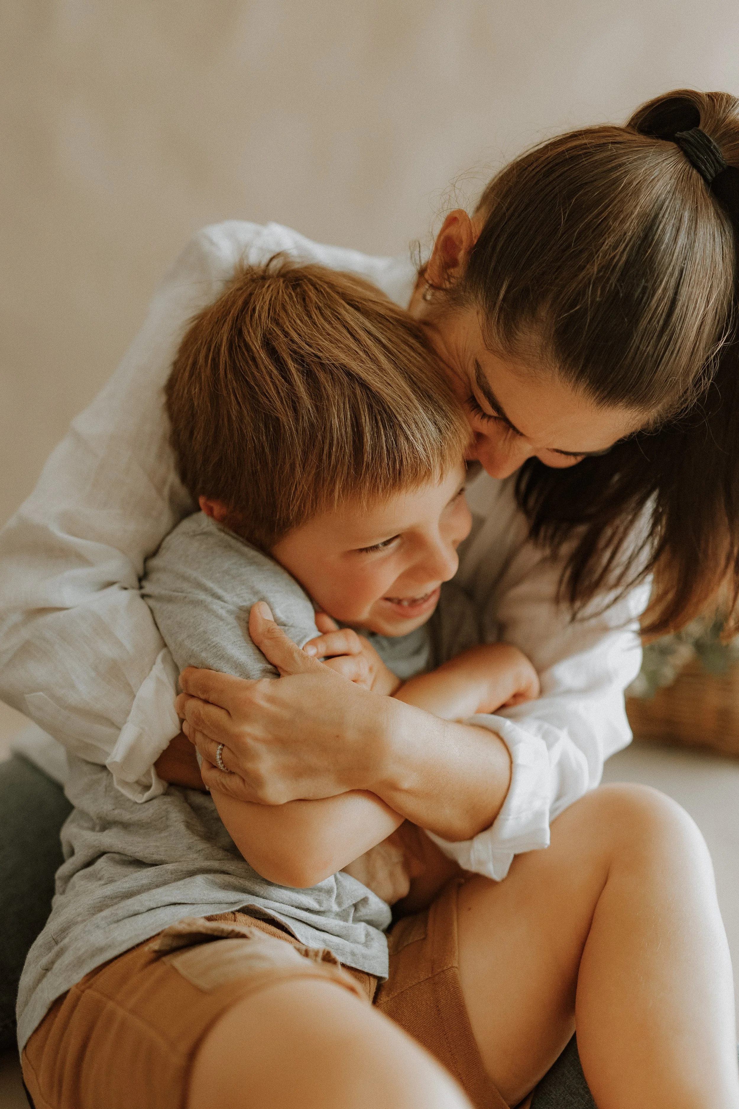 A woman with brown hair in a ponytail is hugging a young boy with light brown hair. They are both smiling and appear to be happy.