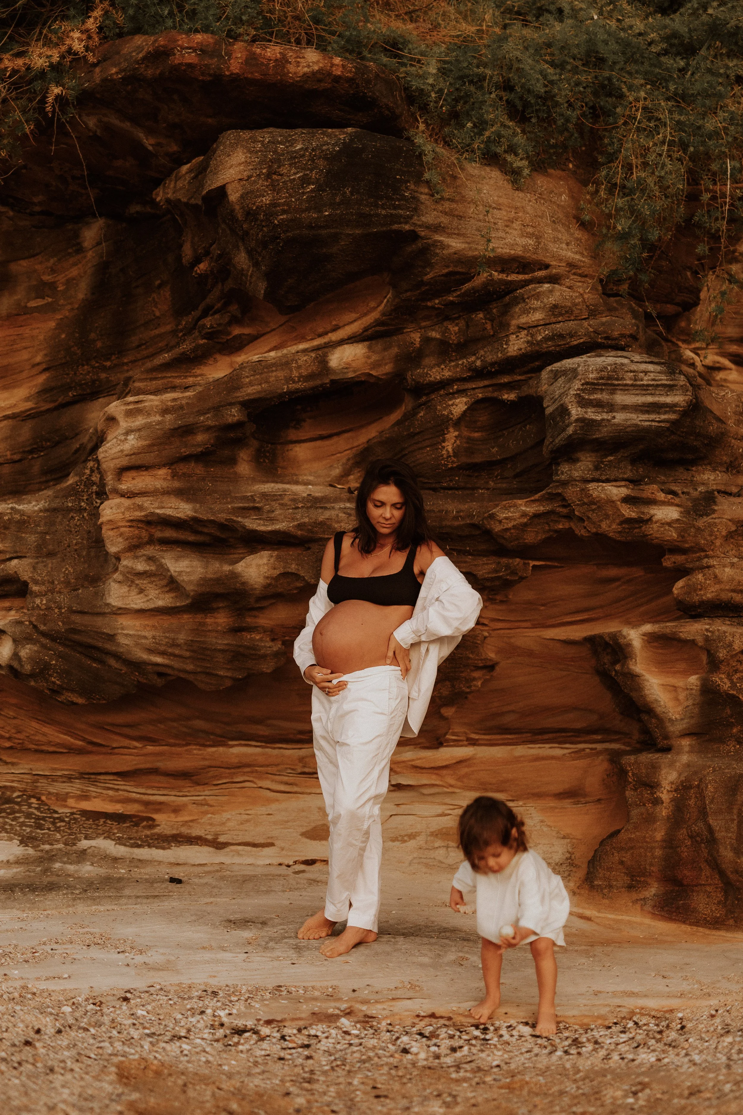 A pregnant woman and two children on a sandy beach with rock formations and green foliage in the background.