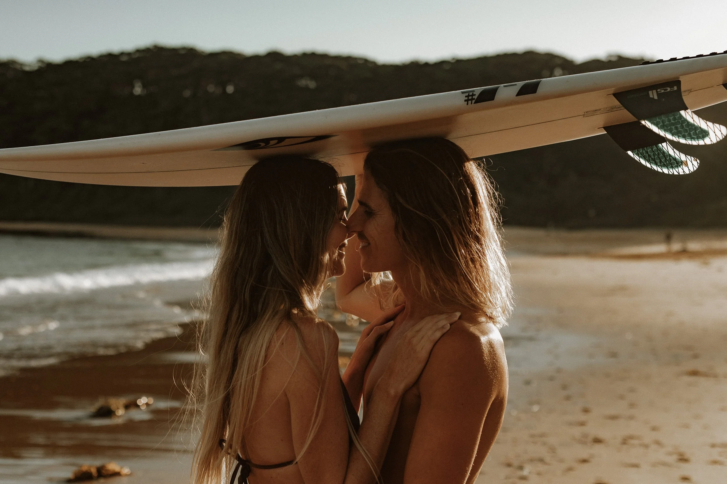 Two women with long hair sharing a close embrace and smiling on a beach at sunset, with a surfboard balanced on their heads.