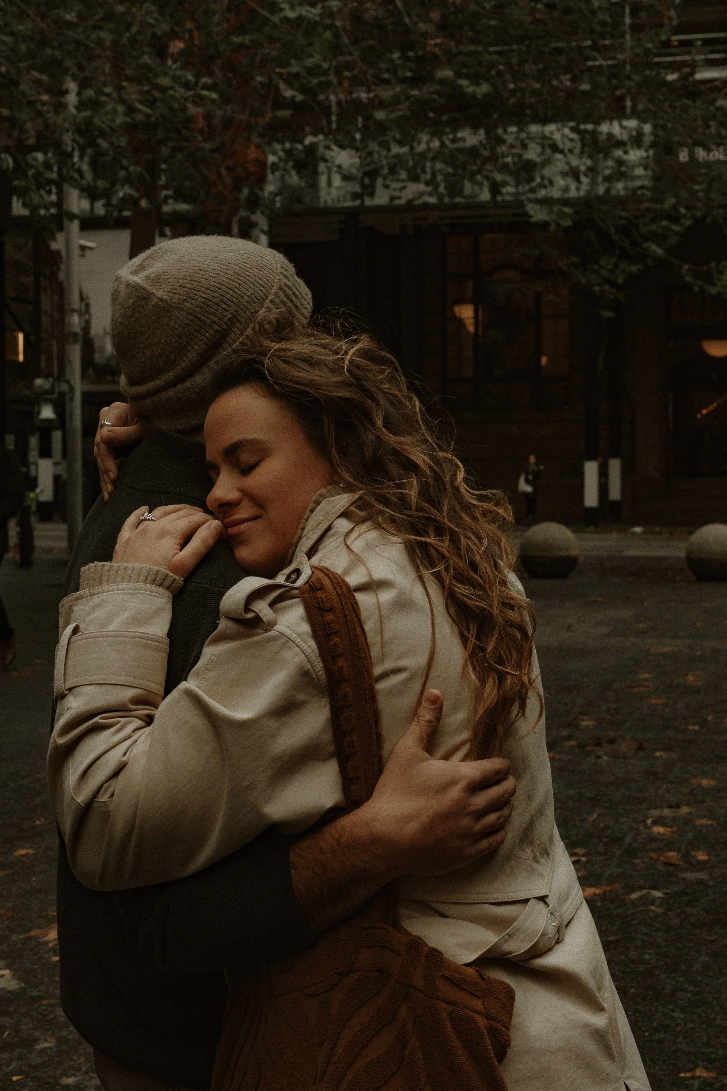 A woman with curly hair hugging a person wearing a beanie, both with eyes closed, embracing warmly outdoors at dusk.