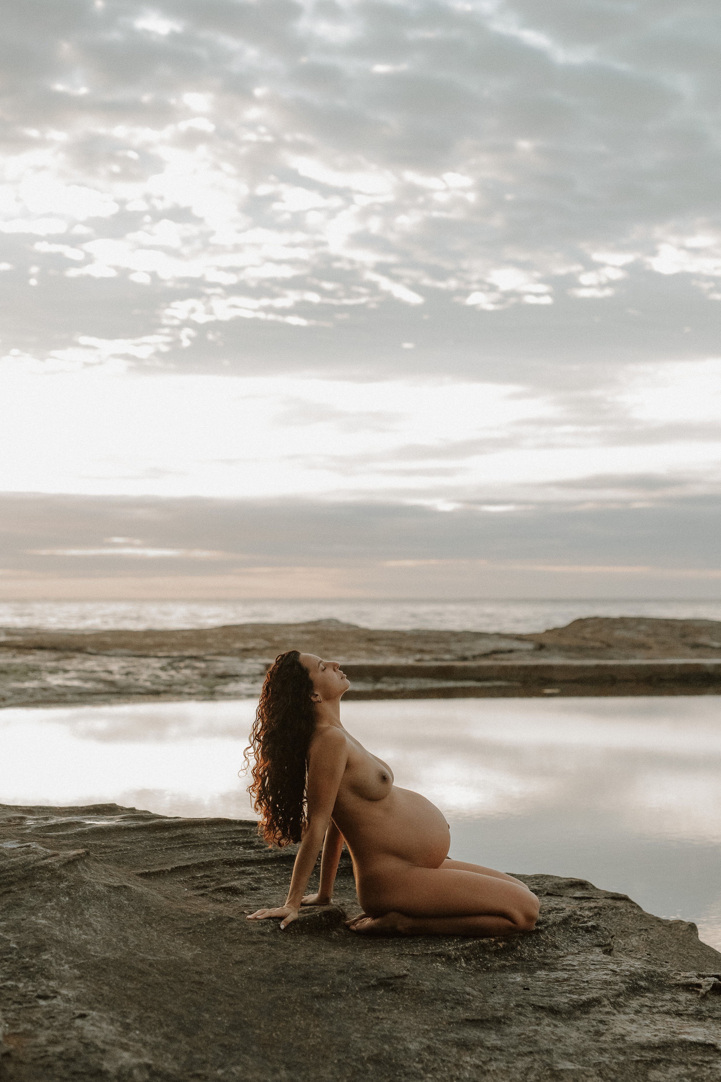 A pregnant woman with long curly hair sitting on a rocky beach with her back arched, looking up at the cloudy sky during sunset.