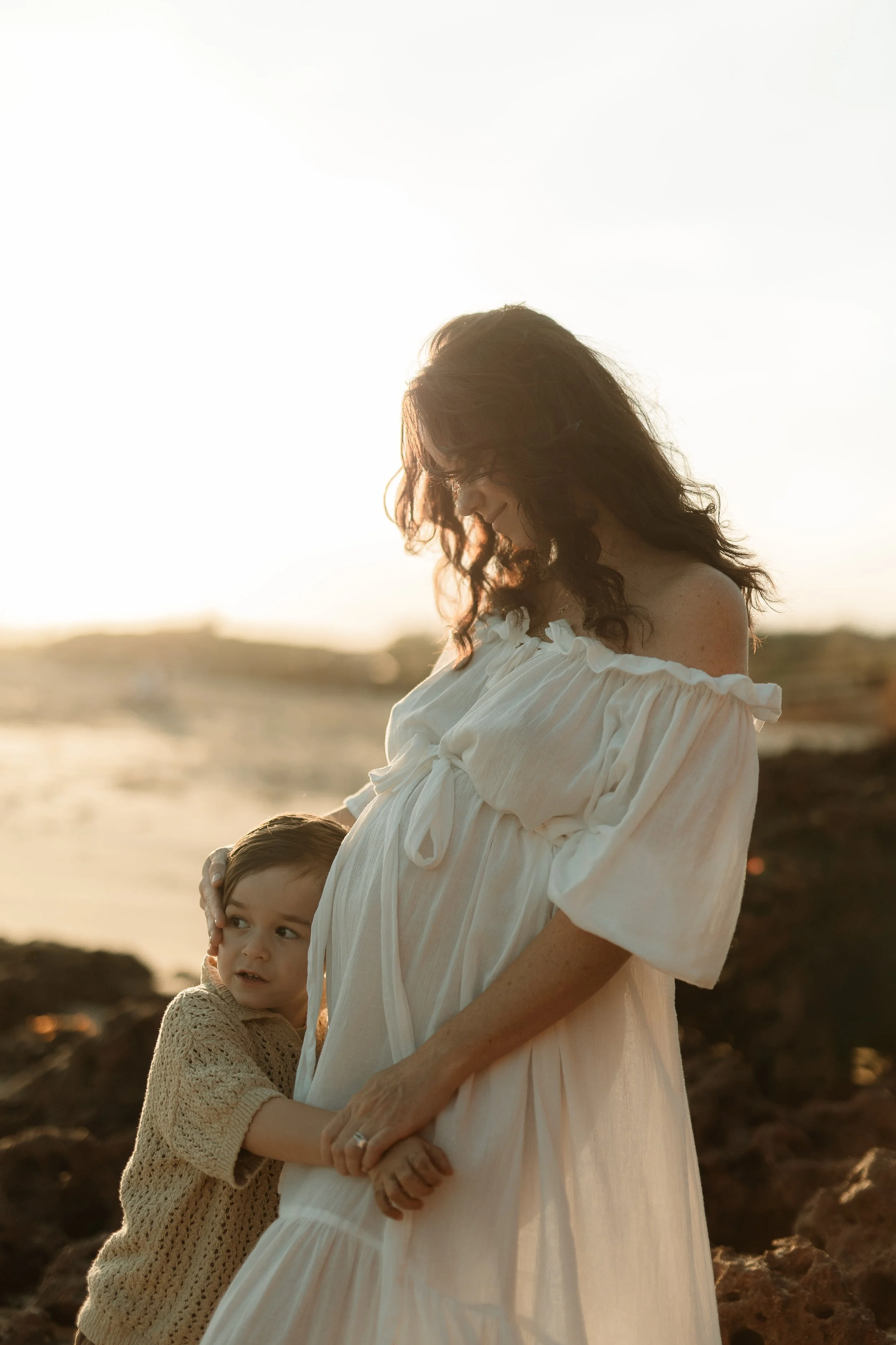 A woman with long, wavy hair and a white, off-the-shoulder dress stands on a rocky beach during sunset, while a young girl with curly hair and a beige sweater holds her hand and looks up at her.