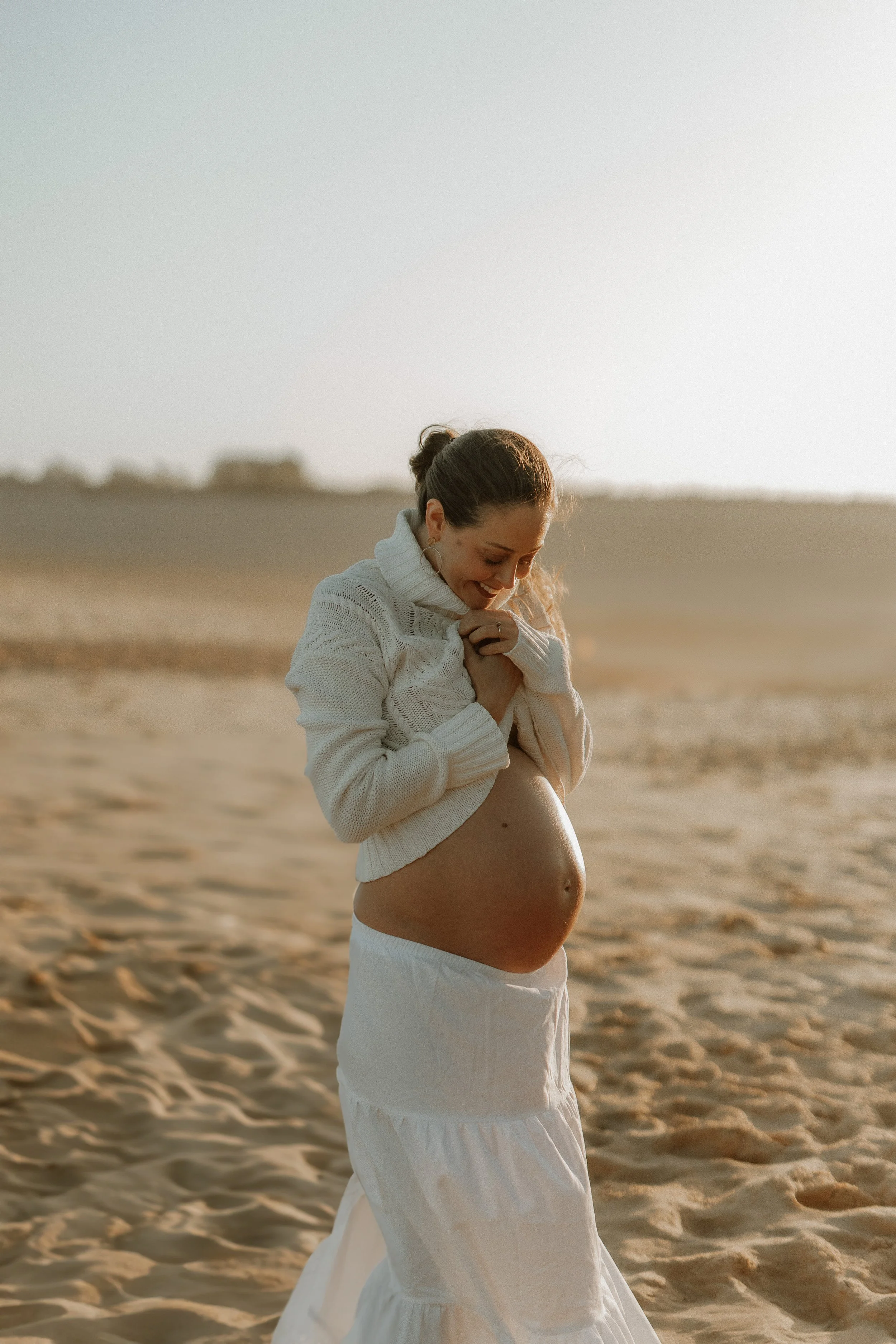A pregnant woman standing on a sandy beach, smiling and looking down with her hands clasped near her chest, wearing a white knitted sweater and a long white skirt, in the golden light of late afternoon.