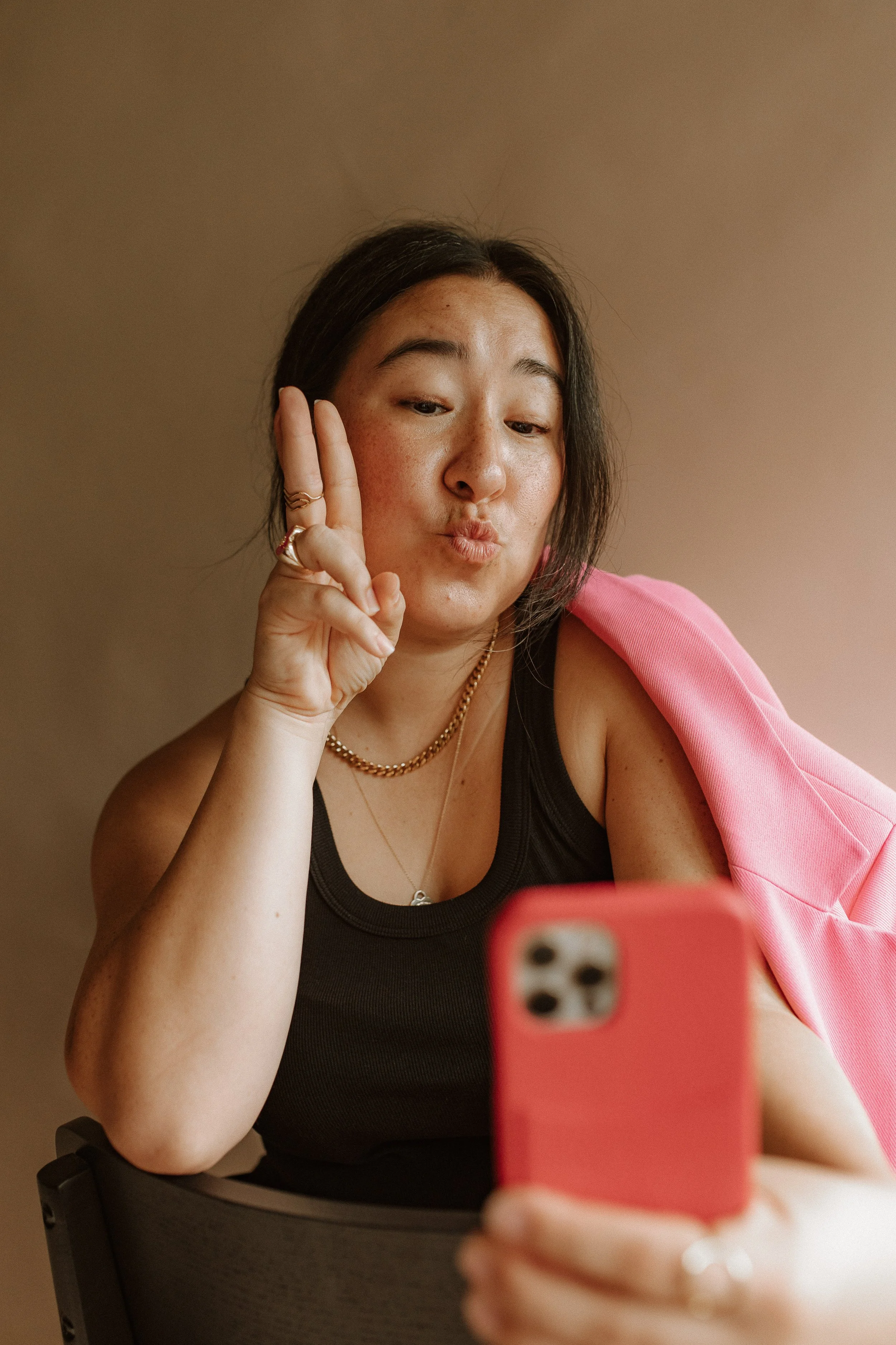 A woman taking a selfie with her smartphone, making a peace sign with her fingers, wearing a black tank top, pink jacket, gold jewelry, and sitting in a chair.