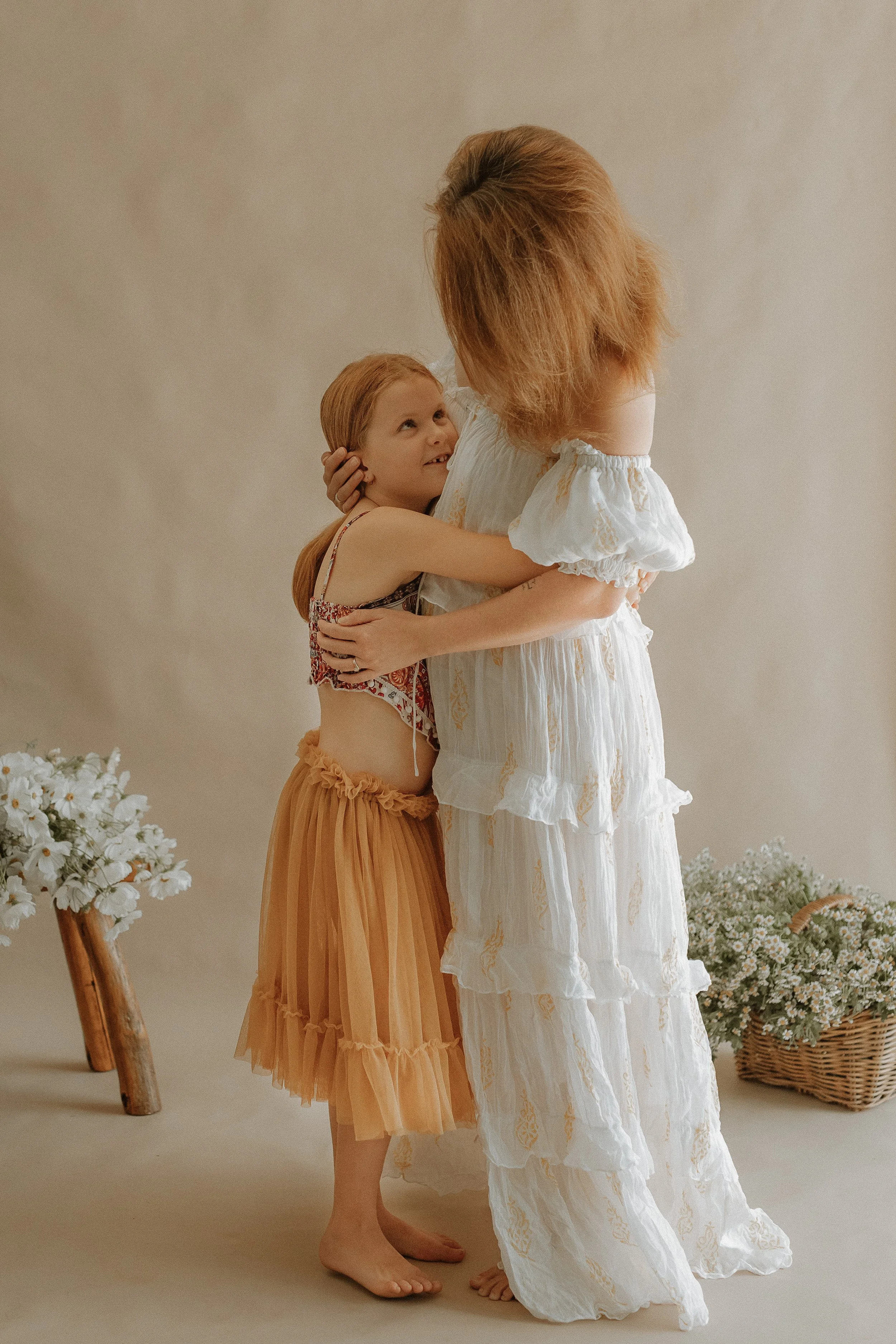 A woman in a white dress hugging a girl with red hair, wearing a patterned tank top and a tan skirt, surrounded by flowers and wicker baskets.