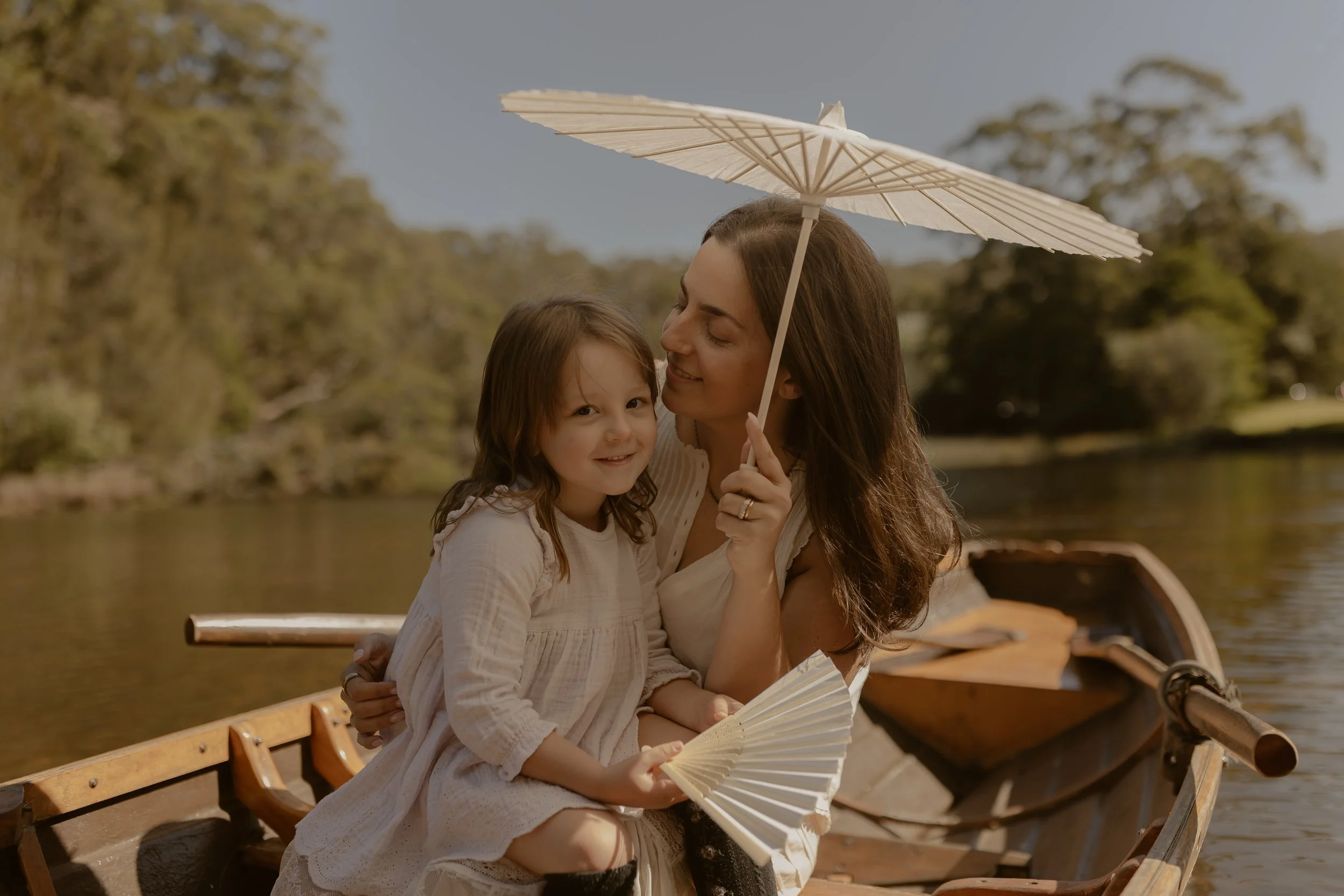 A woman and a young girl sitting in a wooden boat on a lake, sharing a moment under a paper parasol, with trees in the background on a sunny day.