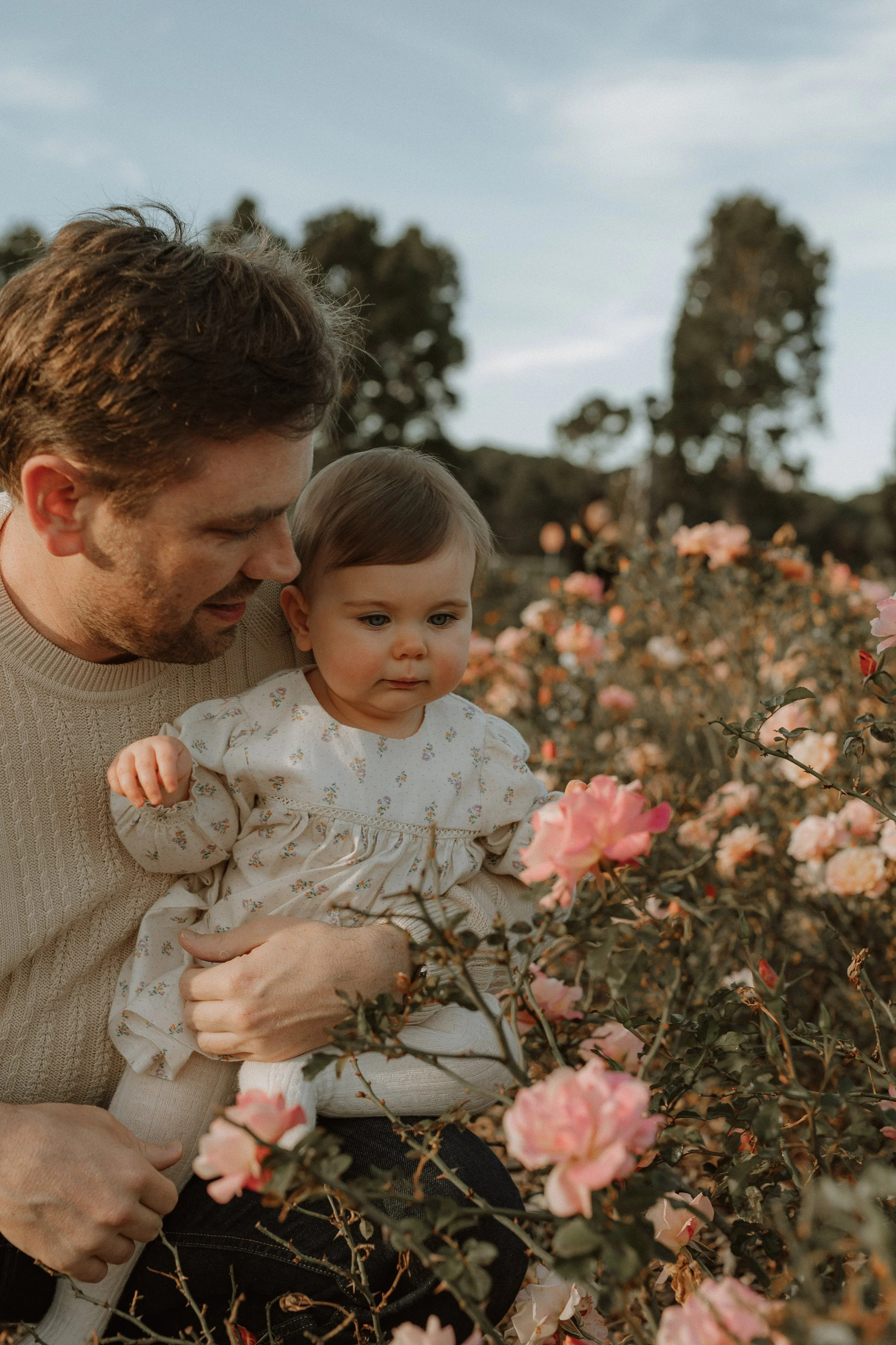 A man holding a young girl in a pink flower garden. The man is smiling and looking at the girl, who is looking at the flowers. The scene takes place outdoors during the day with trees and a cloudy sky in the background.