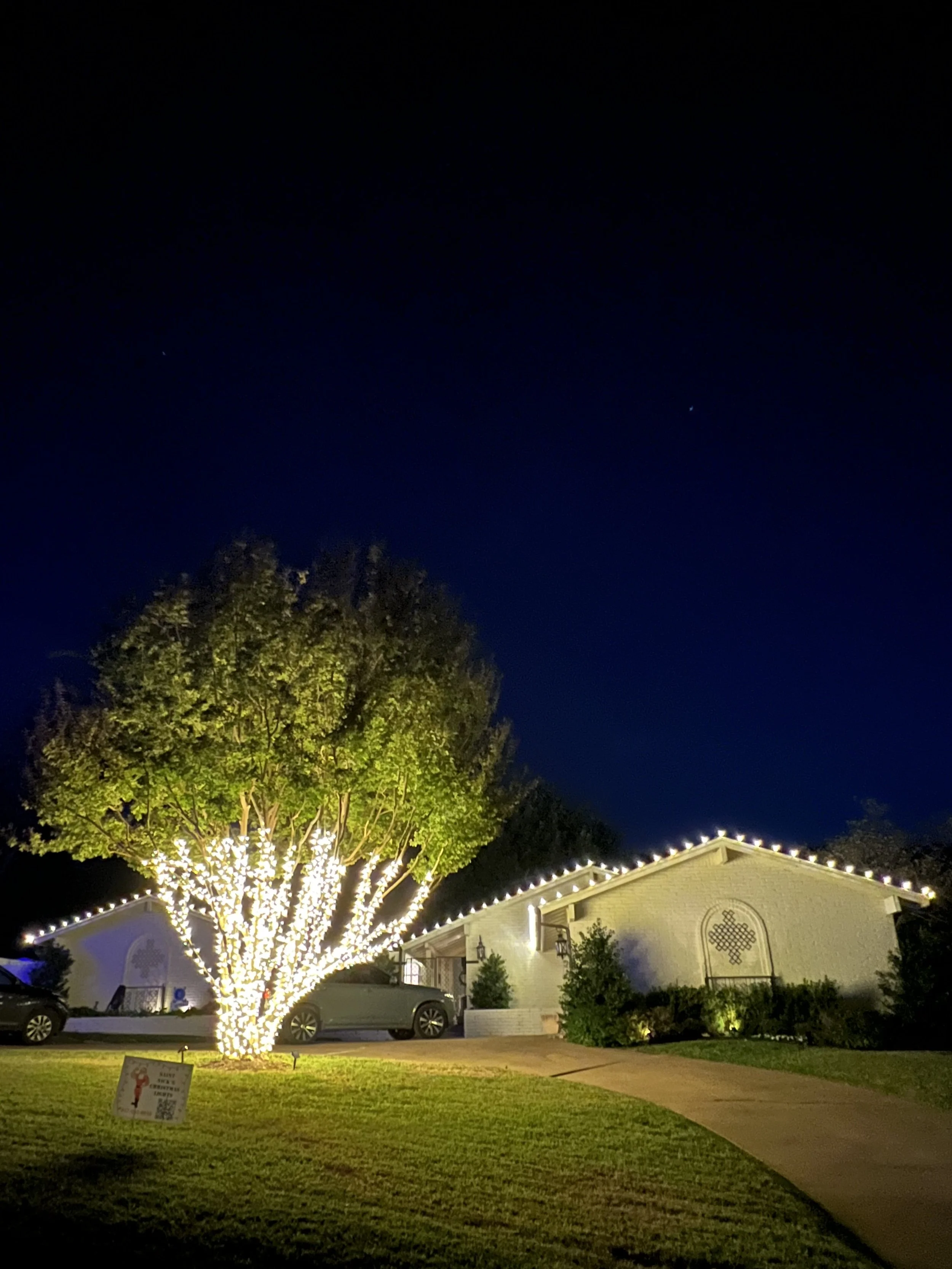 A house decorated with string lights outlining the roof, parked cars, a large lit tree in front, a well-maintained lawn, and a dark night sky with a few visible stars.