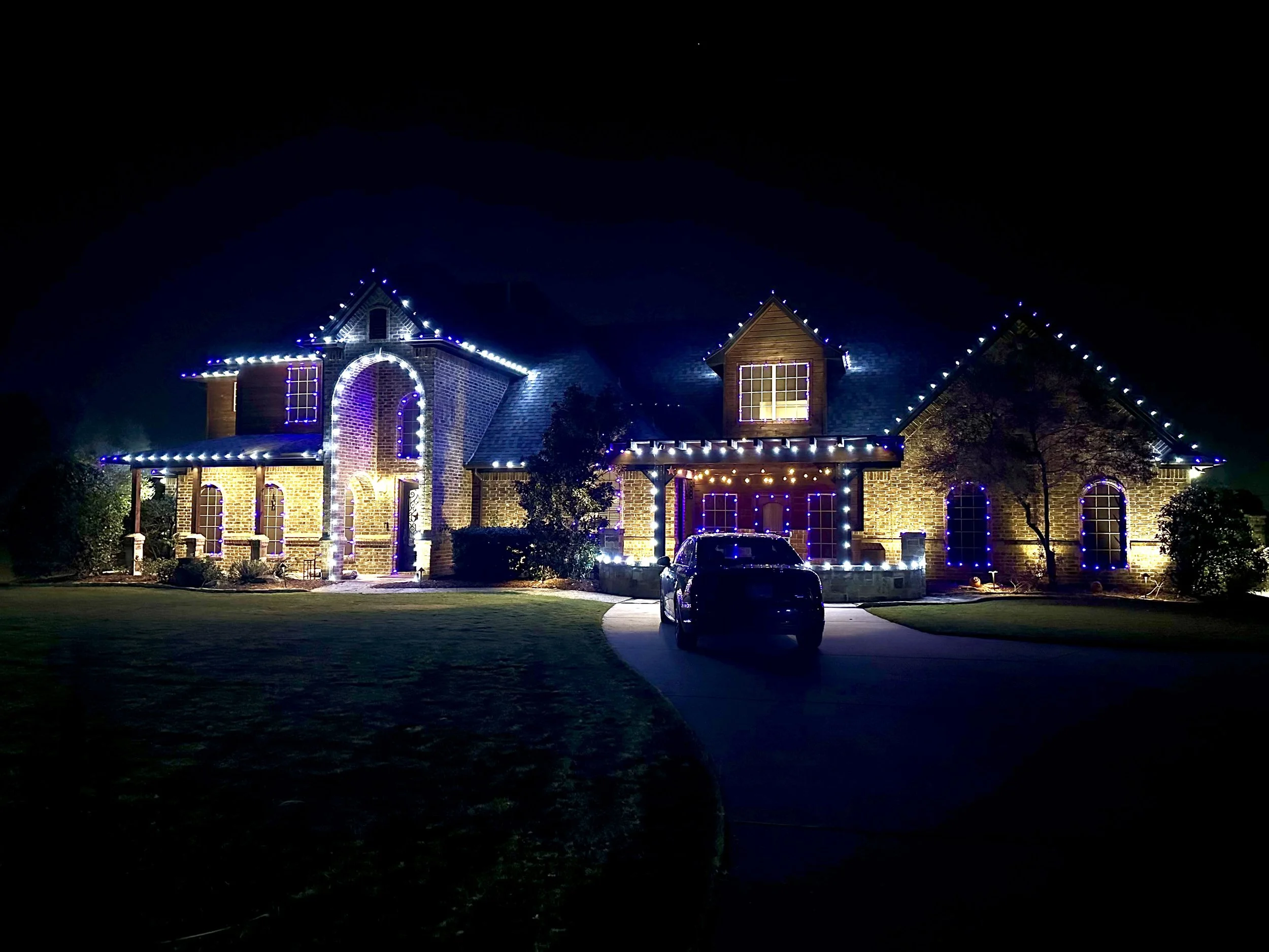 A large brick house decorated with Christmas string lights at night. The house features multiple tiers and windows, with a driveway and a parked car in front.