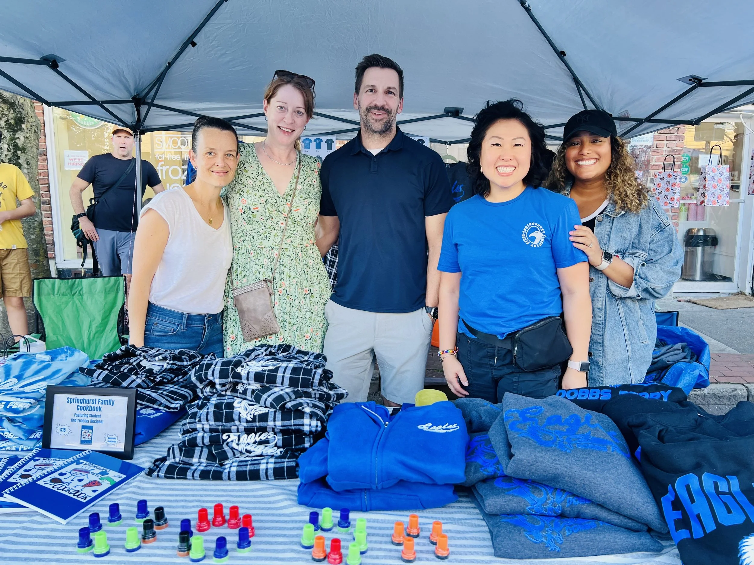 Group of six people standing behind a table with various items at an outdoor event or market, under a blue canopy tent.