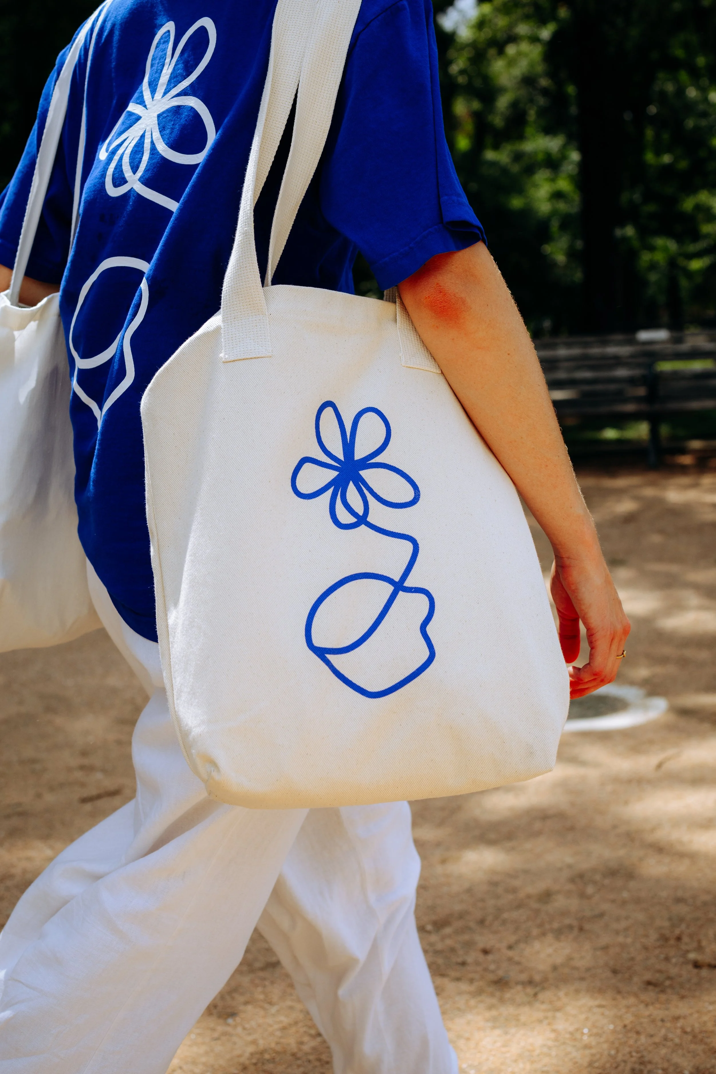A person wearing a blue shirt and white pants carrying a white tote bag with a blue line drawing of a flower and a pot, walking outdoors.