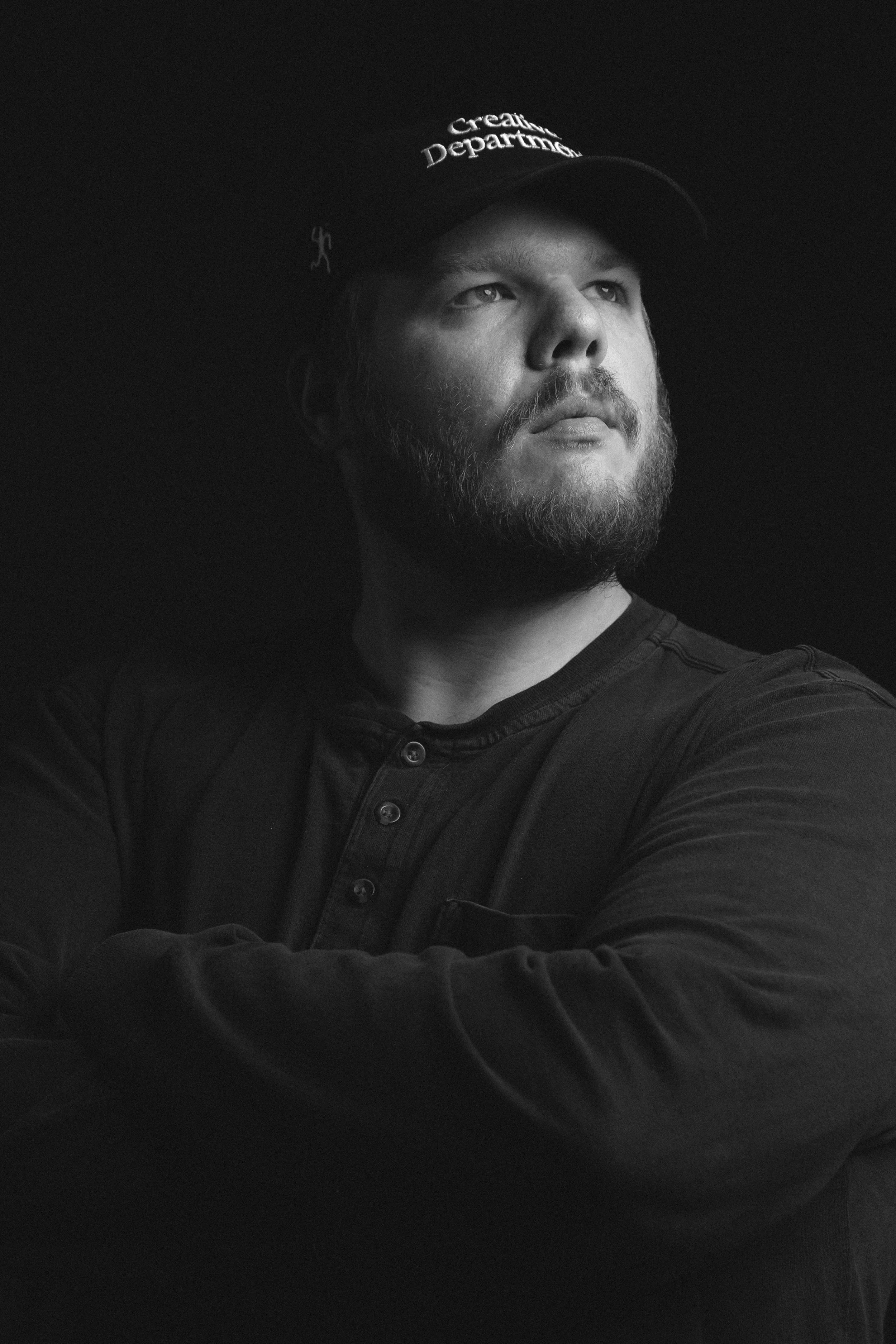 A young man with a beard wearing a black cap and black shirt, looking to his right, against a black background.