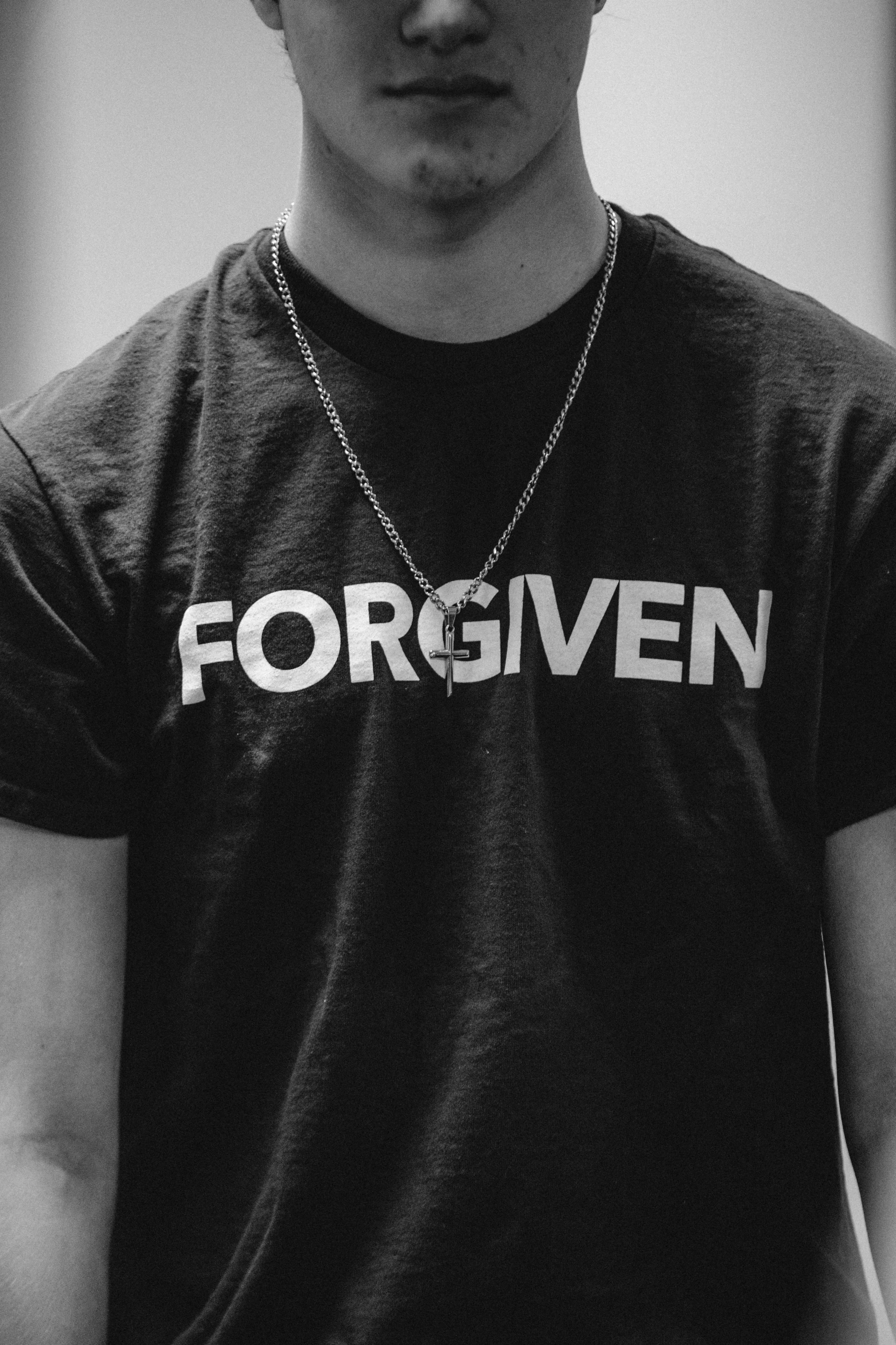 A man wearing a black T-shirt with the word "FORGIVEN" printed across the chest, along with a chain necklace with a cross pendant.