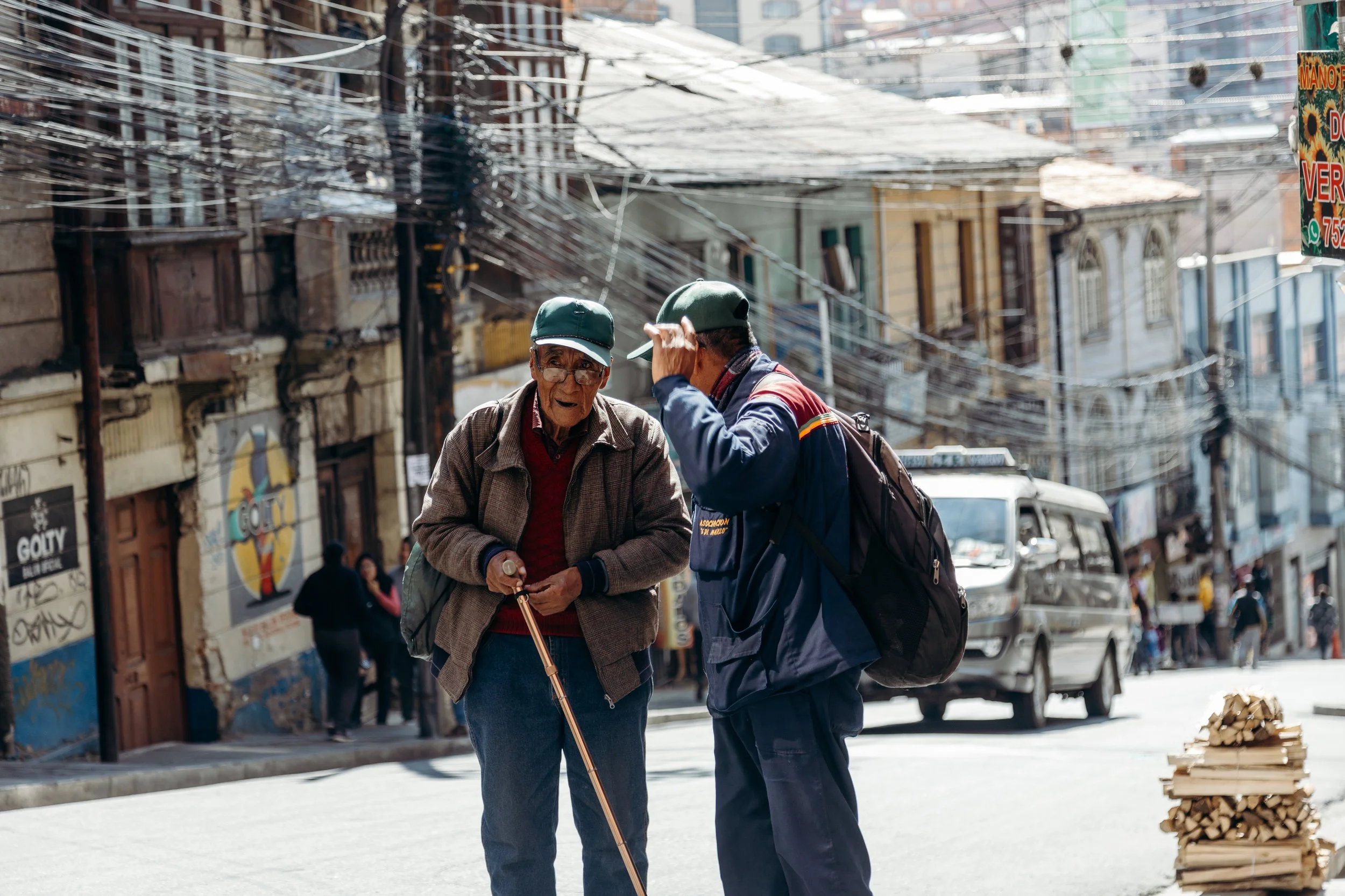 Two elderly men are standing and talking on a busy street with colorful buildings, parked vehicles, and pedestrians in the background. One man is using a cane and wearing a green cap, tan jacket, and glasses, while the other is wearing a green baseba