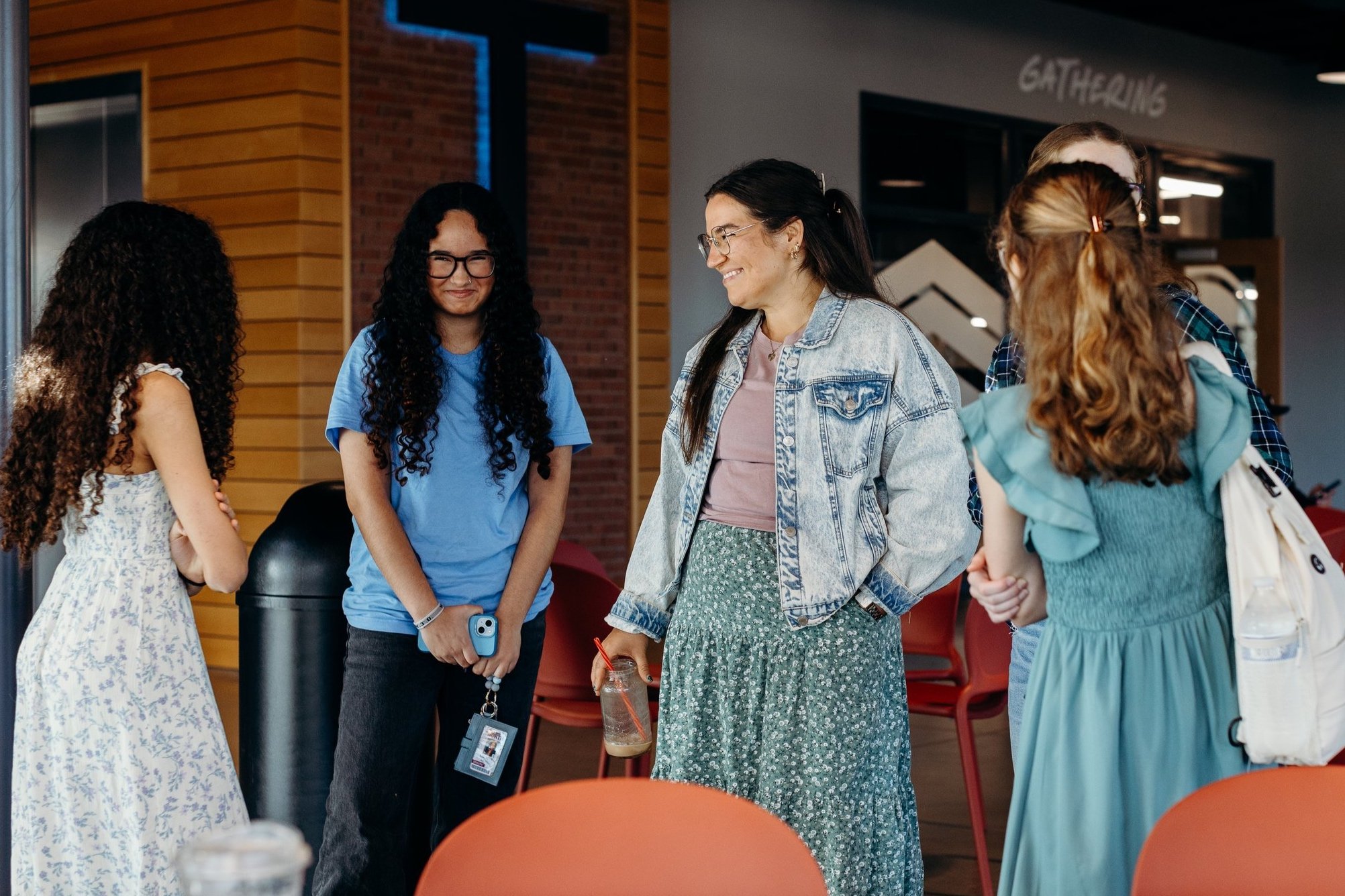 A group of young girls and a woman are talking and smiling indoors, near a sign that says 'Gathering', in a casual setting.