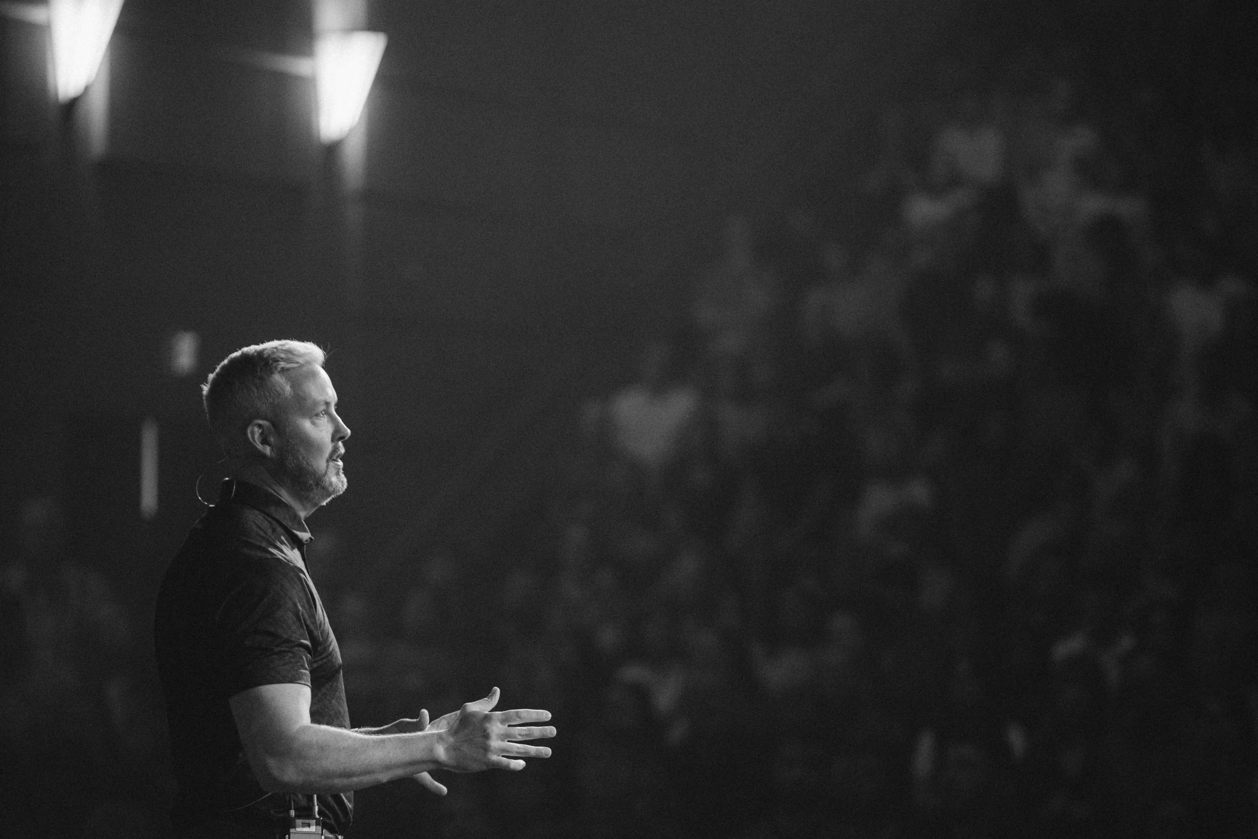 Side profile of a man with short hair and beard speaking on stage in a dark setting.