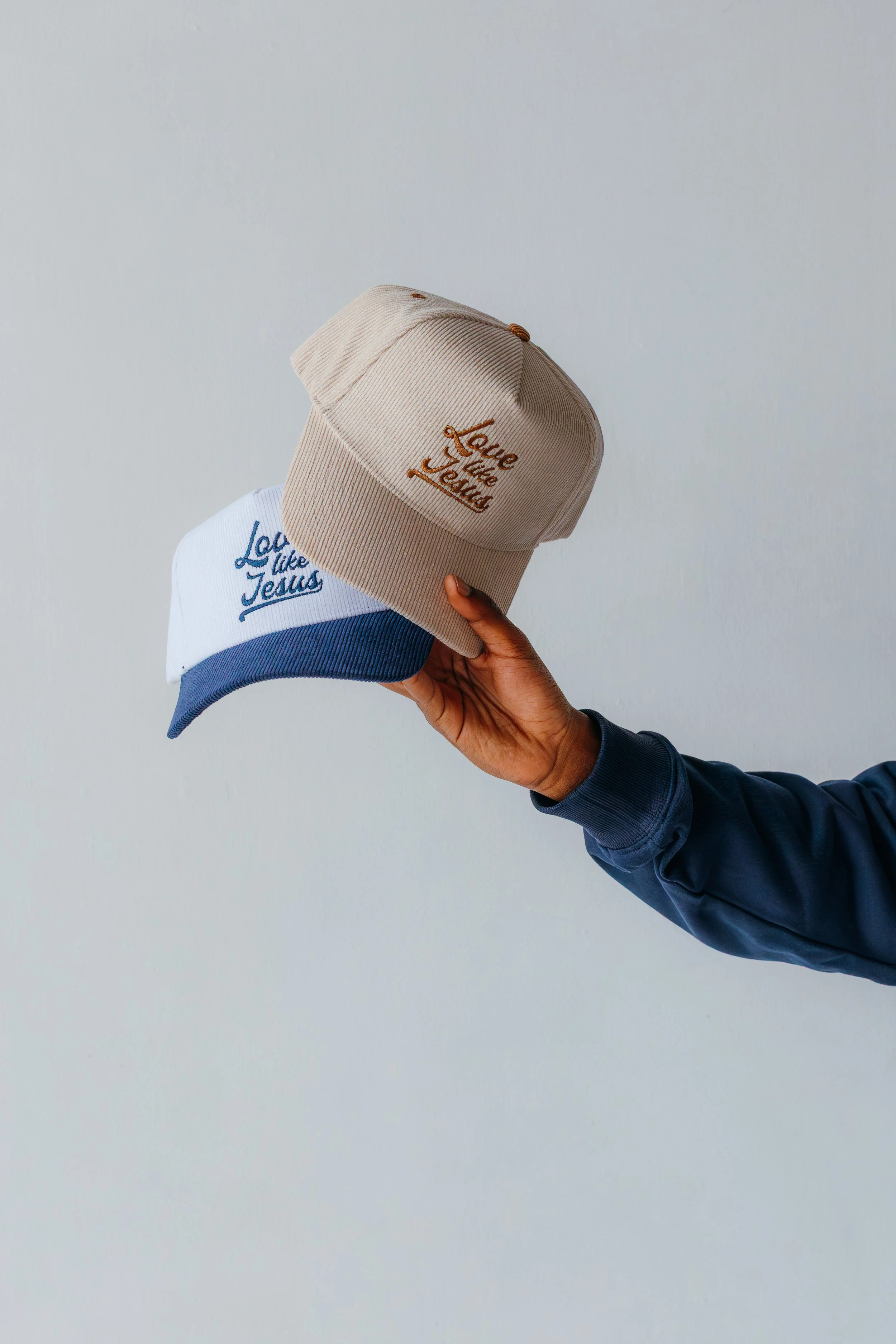 Person holding two baseball caps, one beige with pink embroidery and one white with blue embroidery, against a plain light background.
