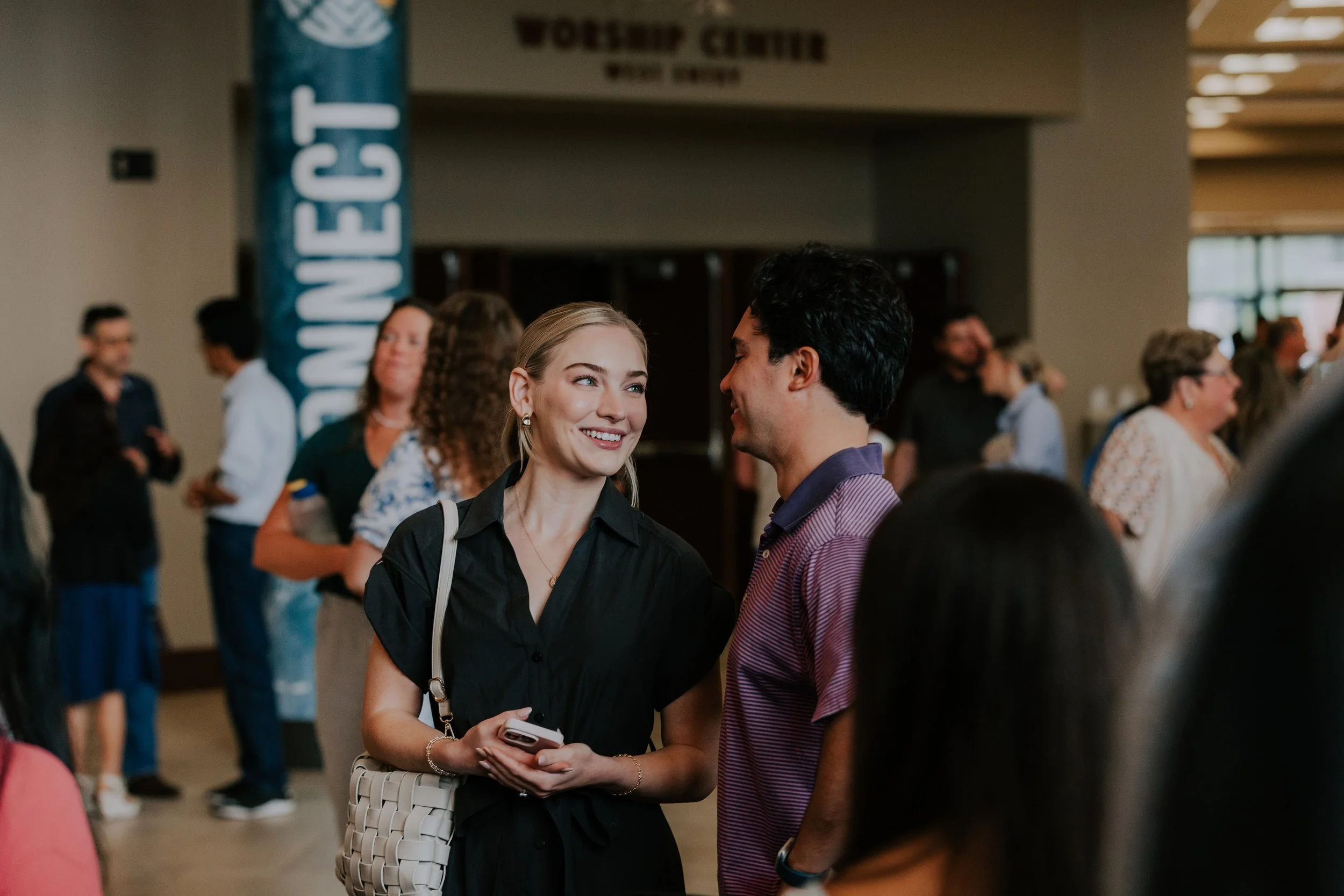 Two people engaged in conversation at a social event with a crowd in the background. The woman has blonde hair, is holding a phone, and is smiling. The man has dark hair and is wearing a striped shirt.