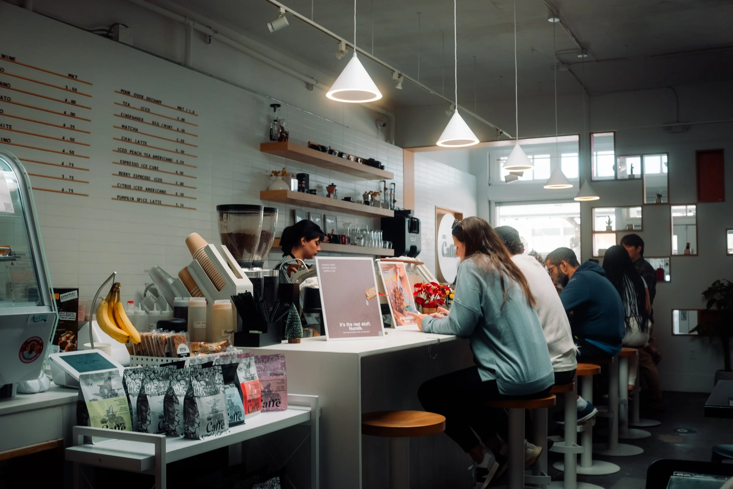 People sitting at a coffee bar inside a cozy cafe, ordering drinks from the barista. The cafe has modern decor with hanging pendant lights, a white tile wall, and shelves with coffee supplies.
