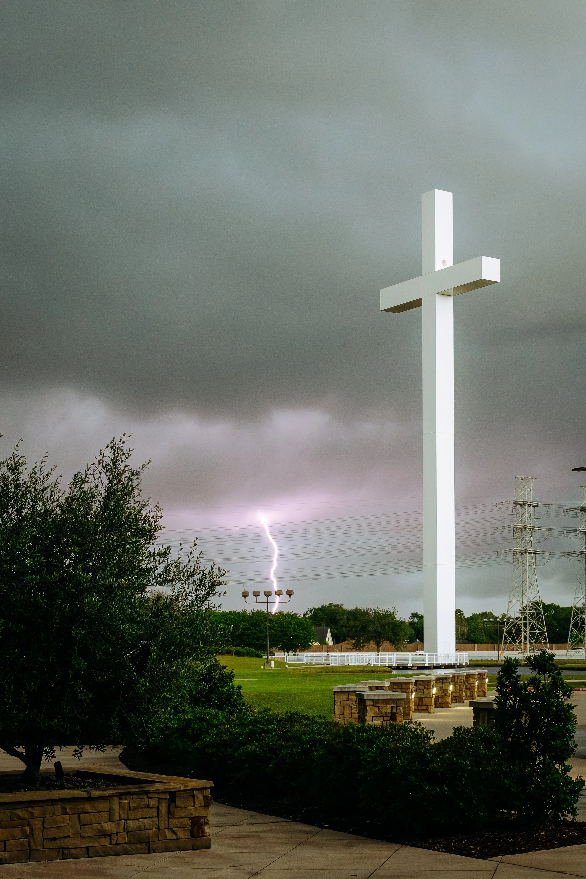 A large white cross structure stands tall against a stormy sky with lightning in the background. The scene includes a park with trees, benches, and a green lawn, with electrical towers and power lines visible in the distance.