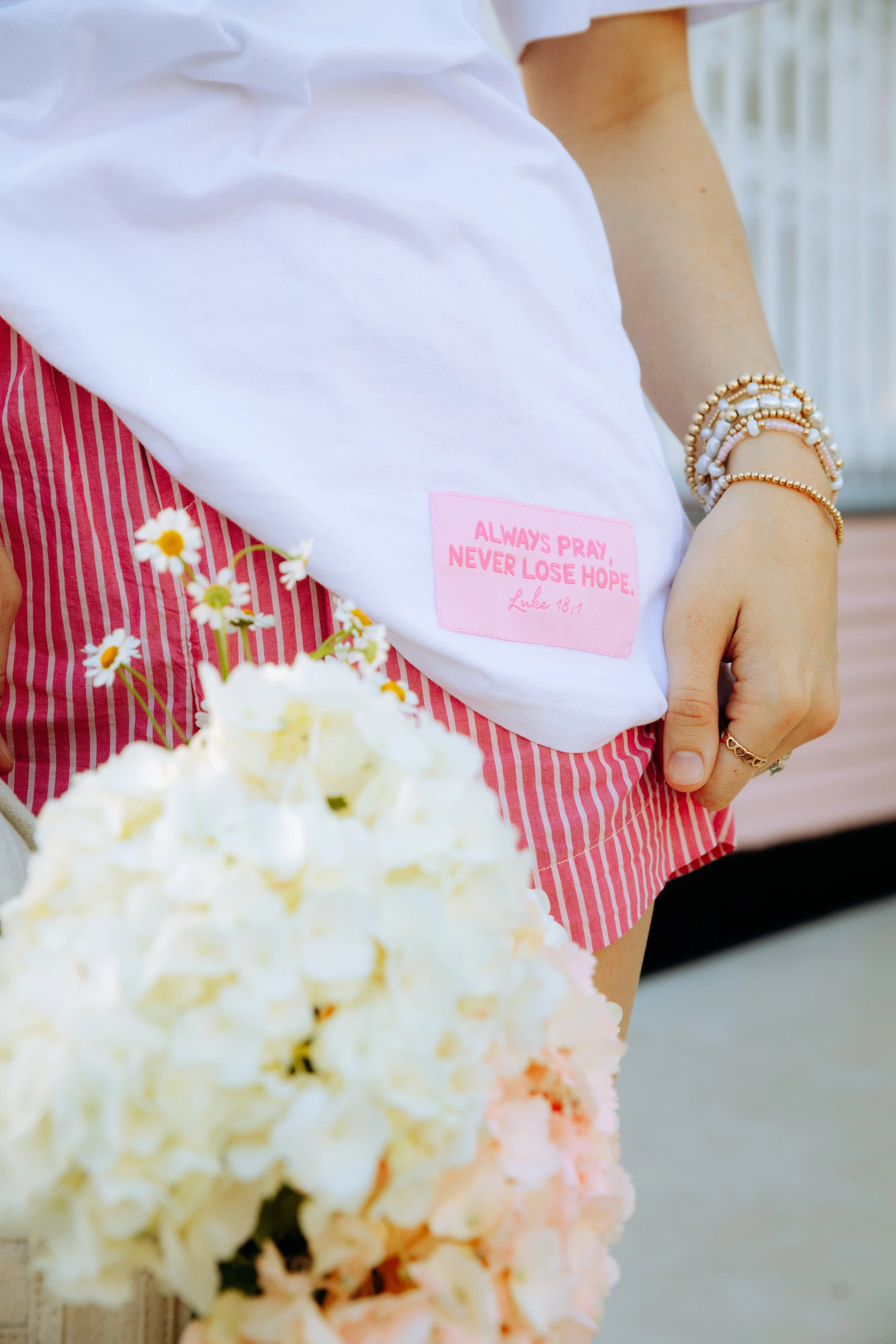 Close-up of a person's white shirt with a pink patch that reads "Always Pray, Never Lose Hope. Luke 18:1," next to a hand with jewelry, and a bouquet of white flowers in the foreground.