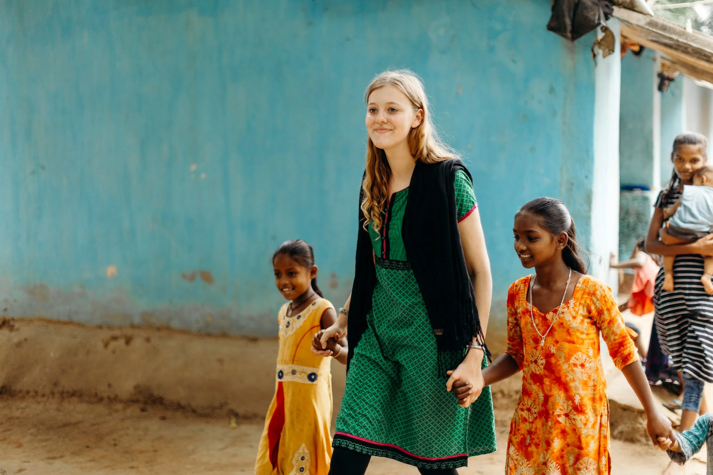 Young woman in green dress holding hands with children, including girls in traditional colorful outfits, walking outdoors near a blue wall.