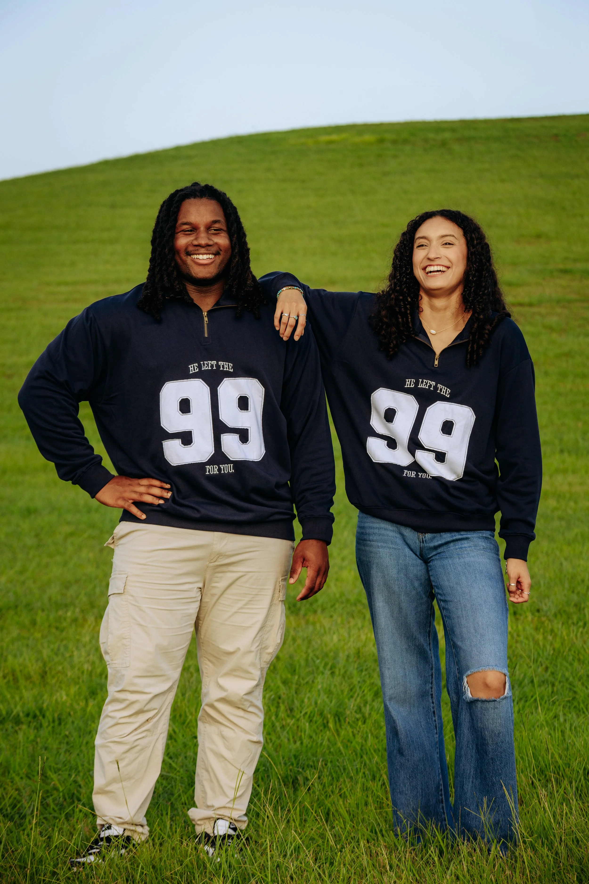 A smiling young man and woman standing on a grassy hill, wearing matching navy blue sweatshirts with the number 99, and the text "He left the 99 for you." The man is wearing beige cargo pants, and the woman is wearing ripped jeans. The woman has her 