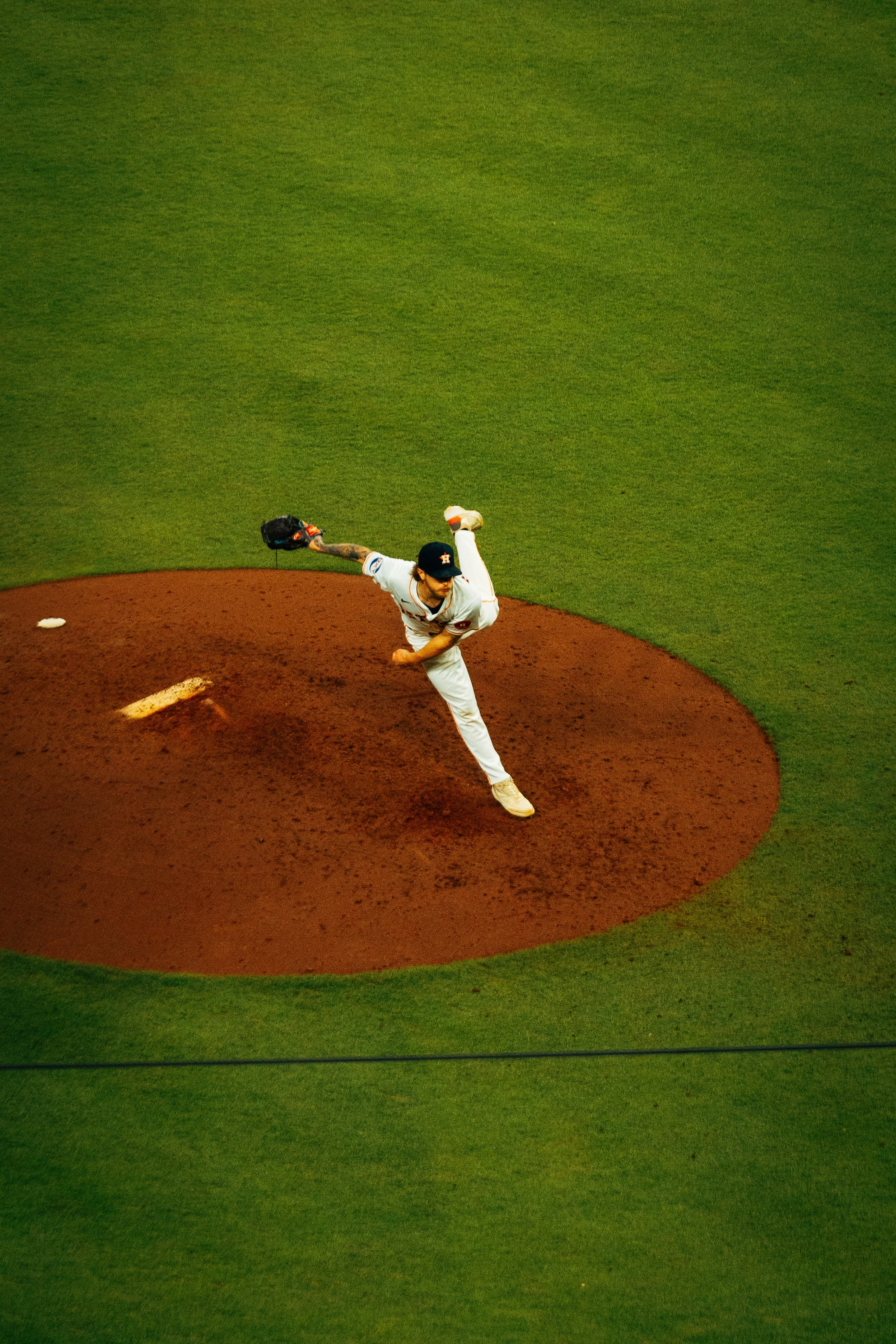 A baseball pitcher in a white uniform is throwing a pitch on a baseball field, viewed from above.