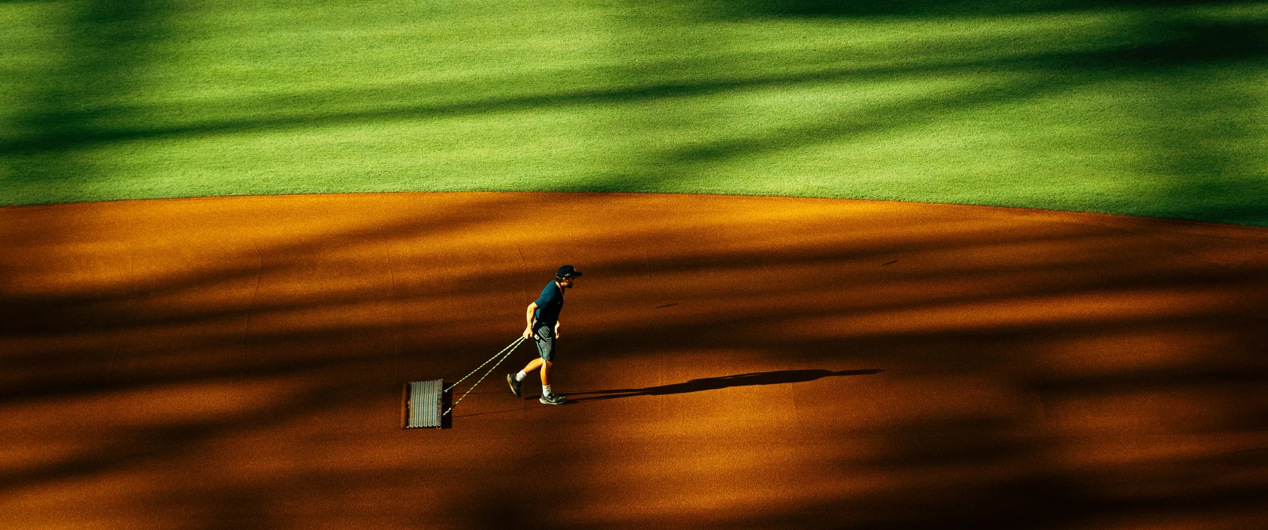A person walking on a baseball or softball infield, pulling a roller behind them, with a grassy outfield in the background.
