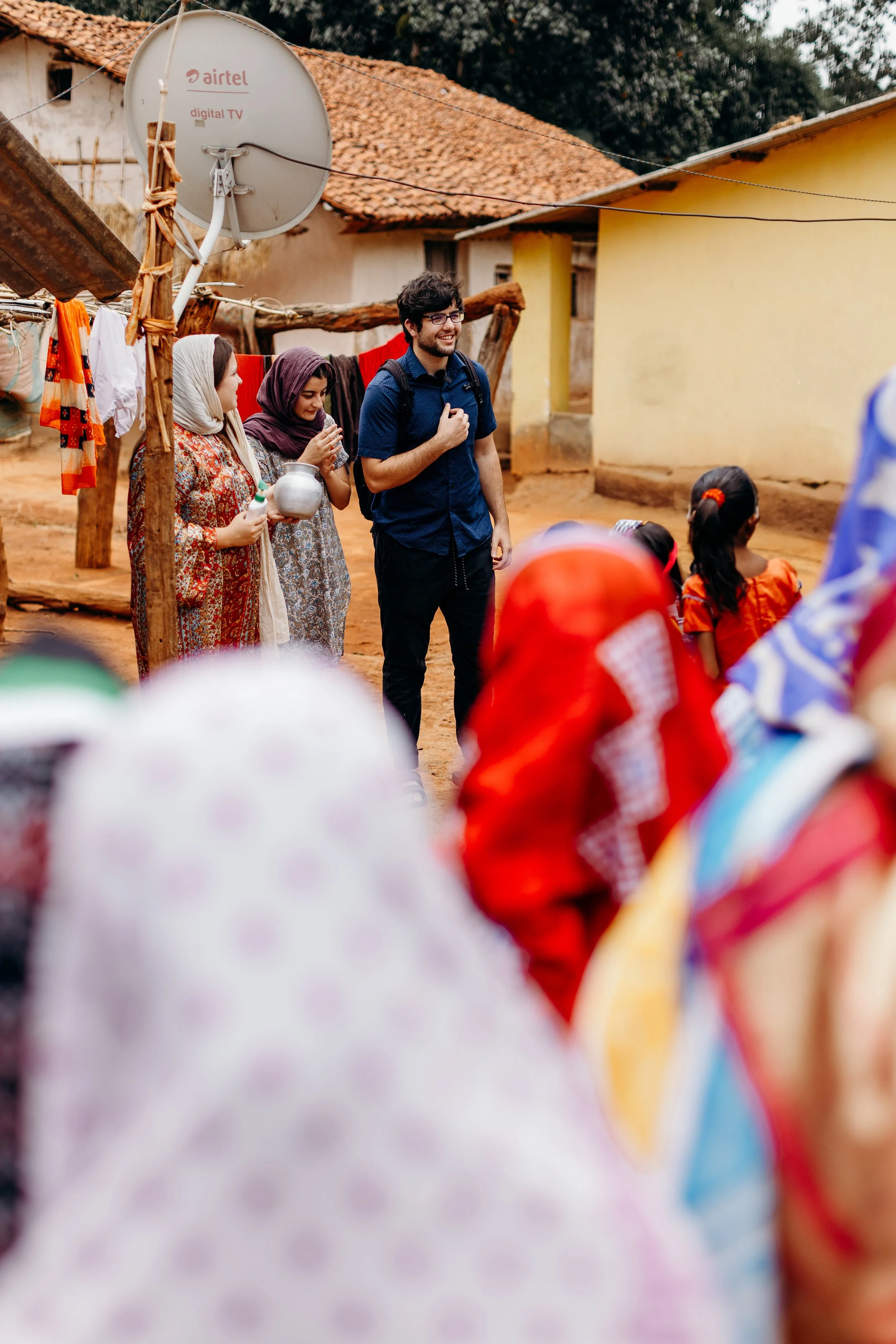 A group of women and children in colorful traditional attire gather outdoors, observing a man in a blue shirt with a backpack, in a rural village setting with houses and a large satellite dish in the background.