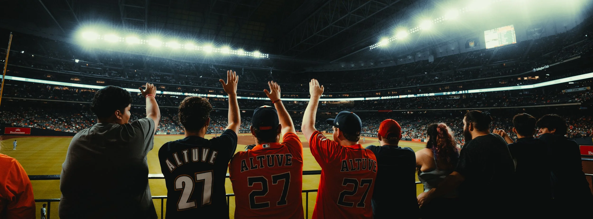 People watching a baseball game at a stadium, some wearing red jerseys with the name 'ALTUVE' and number 27, with bright stadium lights overhead.