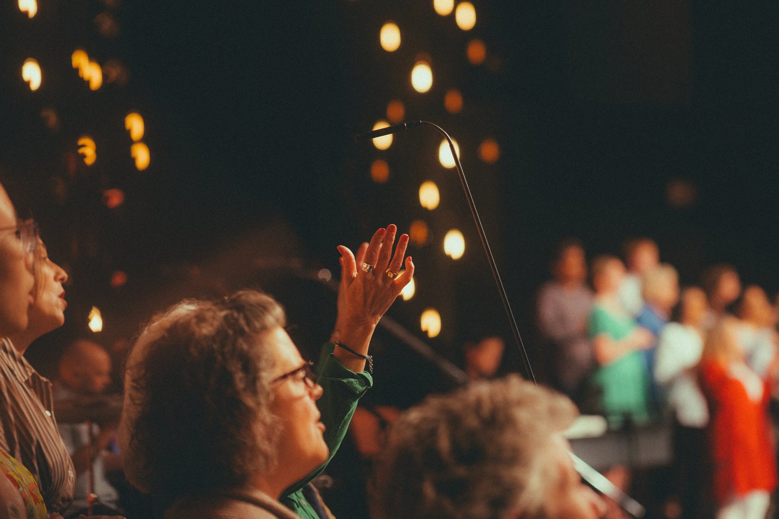A woman raising her hand during an indoor event with a dark background and hanging lights.
