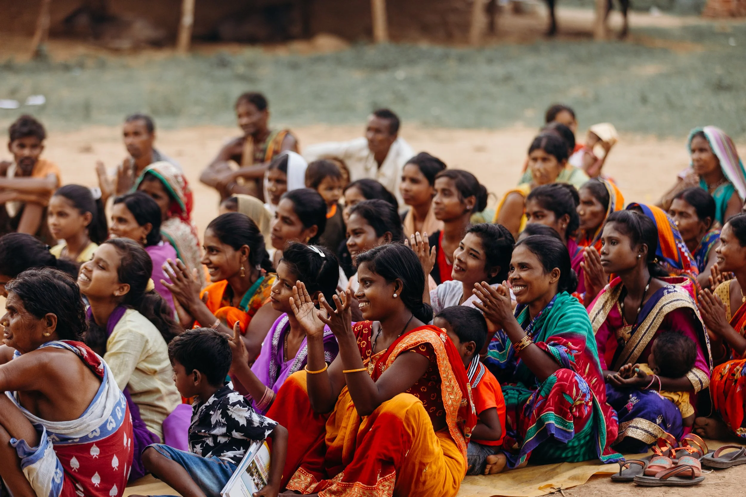 A large group of women and children sitting on the ground, smiling and clapping, dressed in colorful traditional clothing.