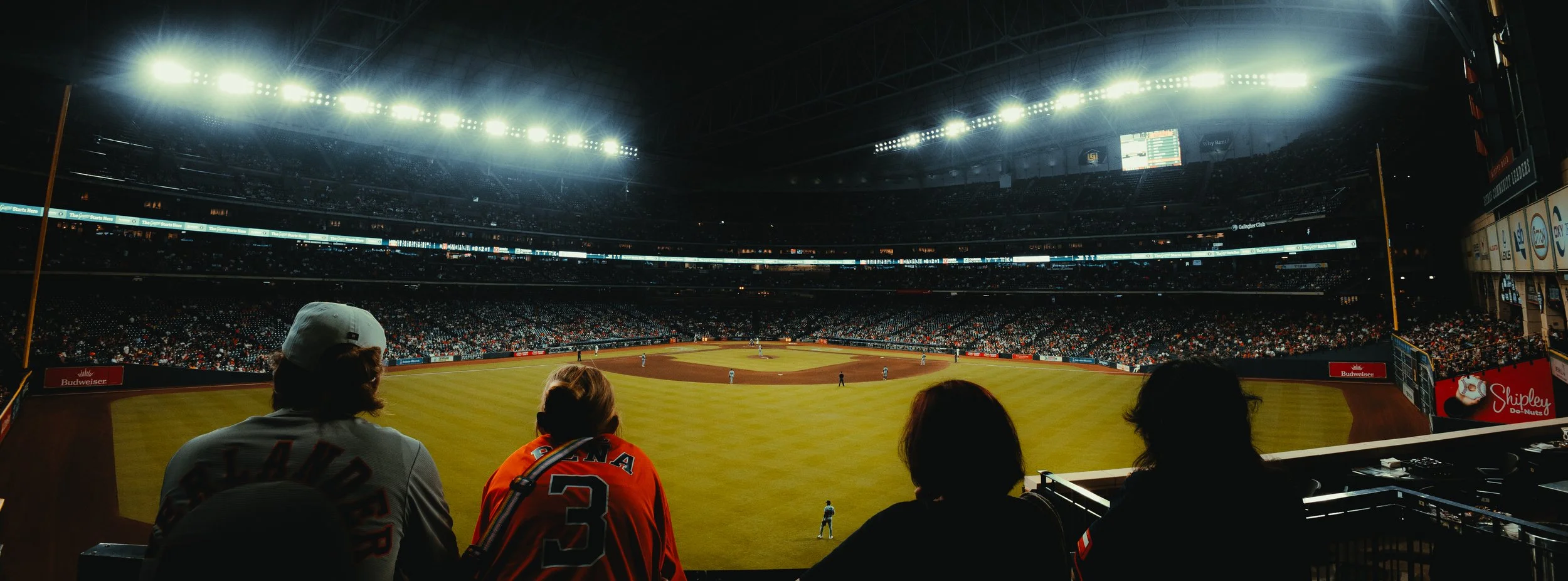 View of a baseball stadium from the stands with fans watching the game, illuminated by bright stadium lights.