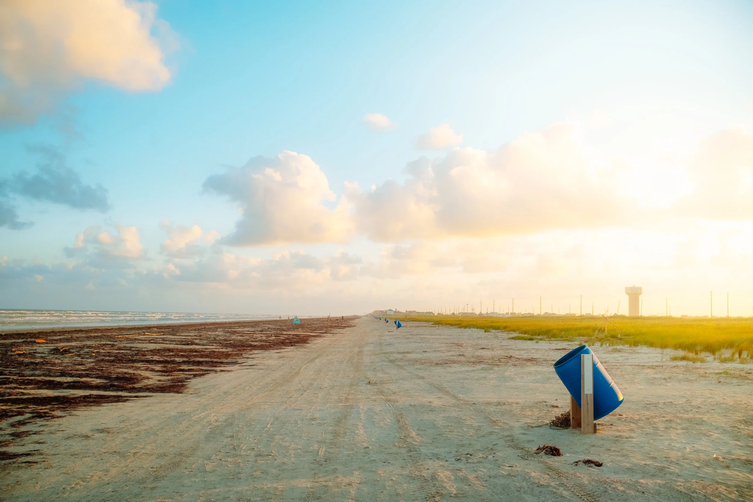 Empty beach with blue trash can tilted on its side, sandy pathway, green grass, distant water, clouds, and a sky with sunlight.