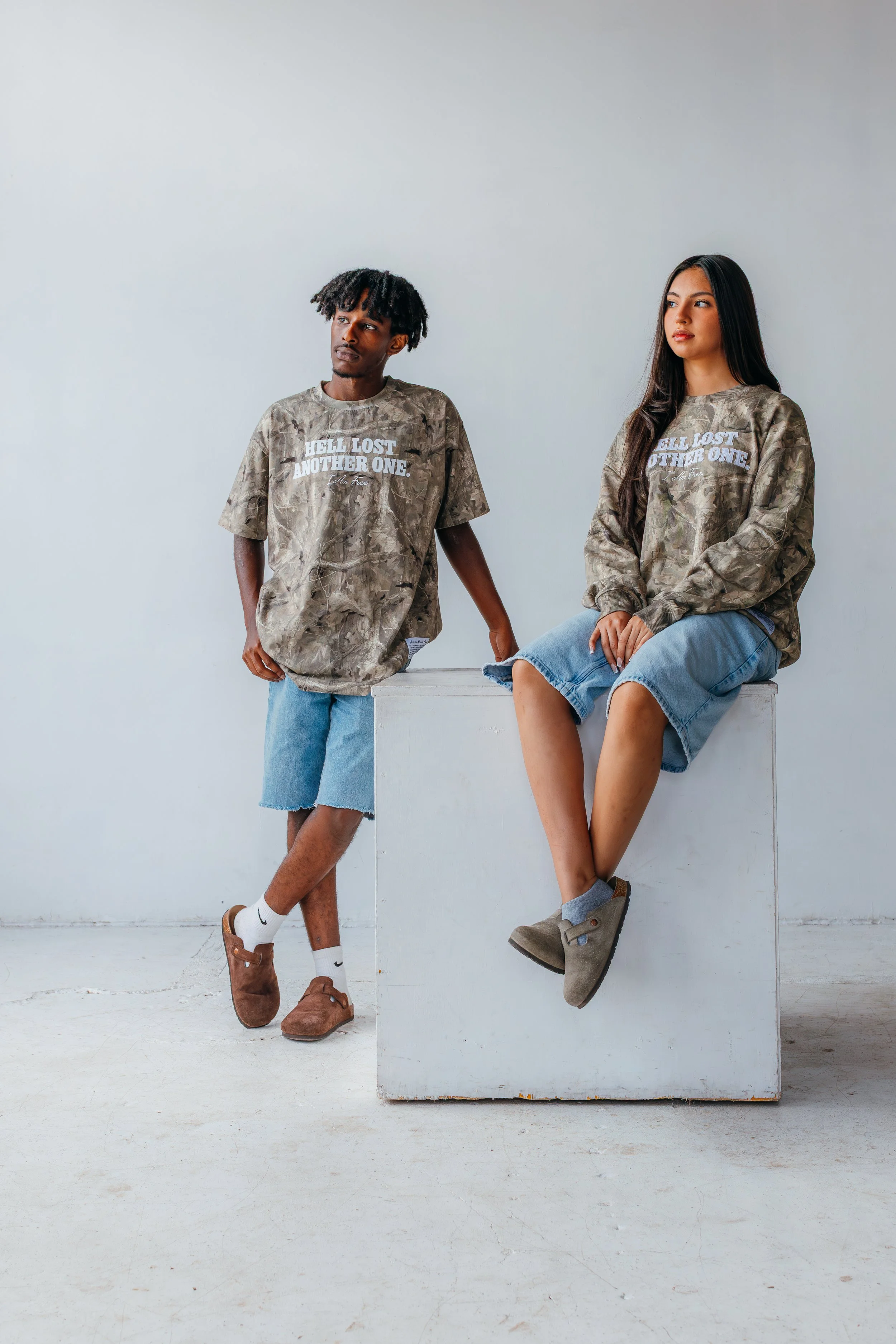 Two young people in casual clothing, one standing and the other sitting on a white box, both wearing matching camouflage-style sweatshirts with the text "Hell Lost, Another One" printed on them.