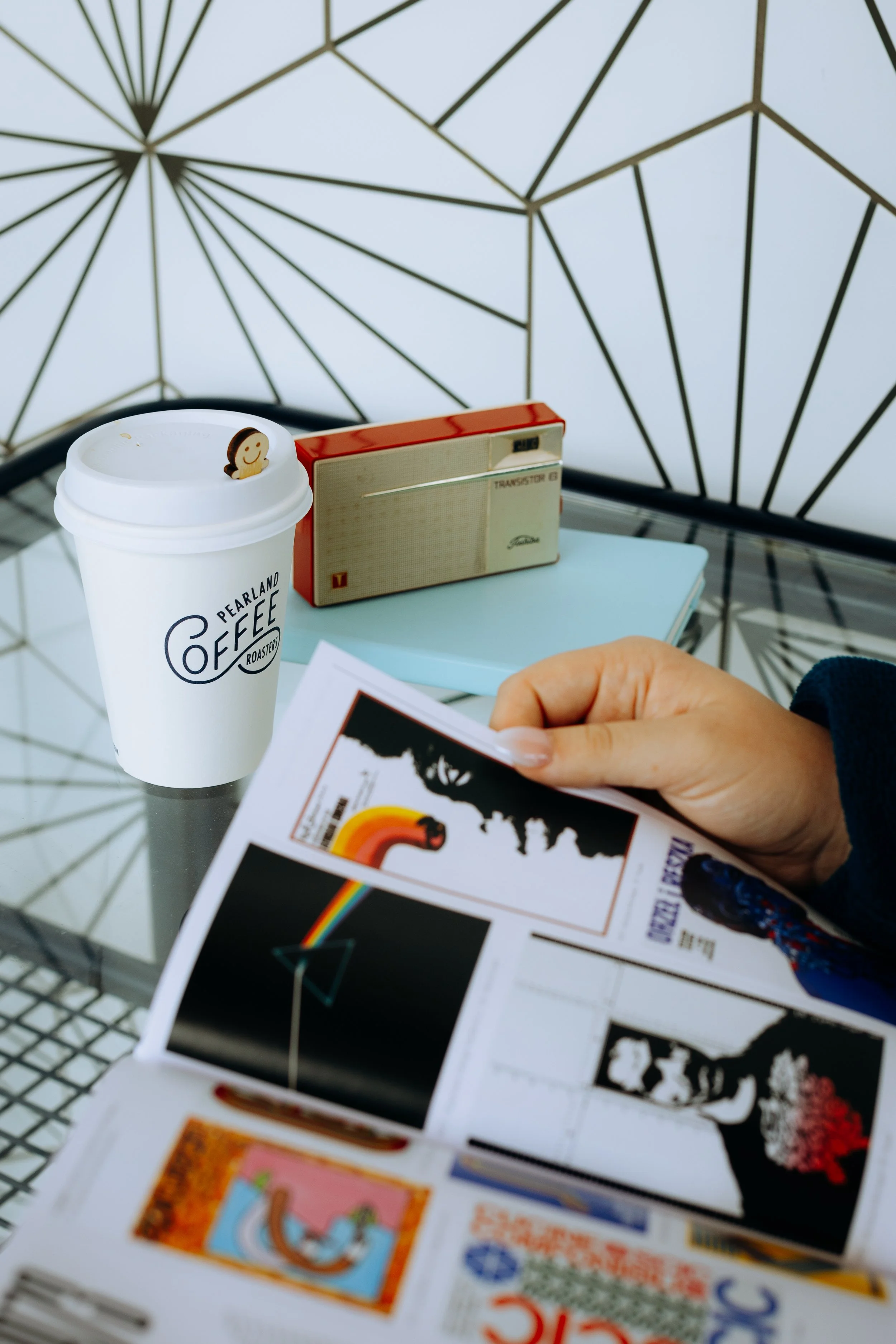 A person holding a colorful sheet of paper with artwork and text, sitting at a glass table with a coffee cup, a vintage transistor radio, and a small blue notebook, with a decorative metal wall in the background.