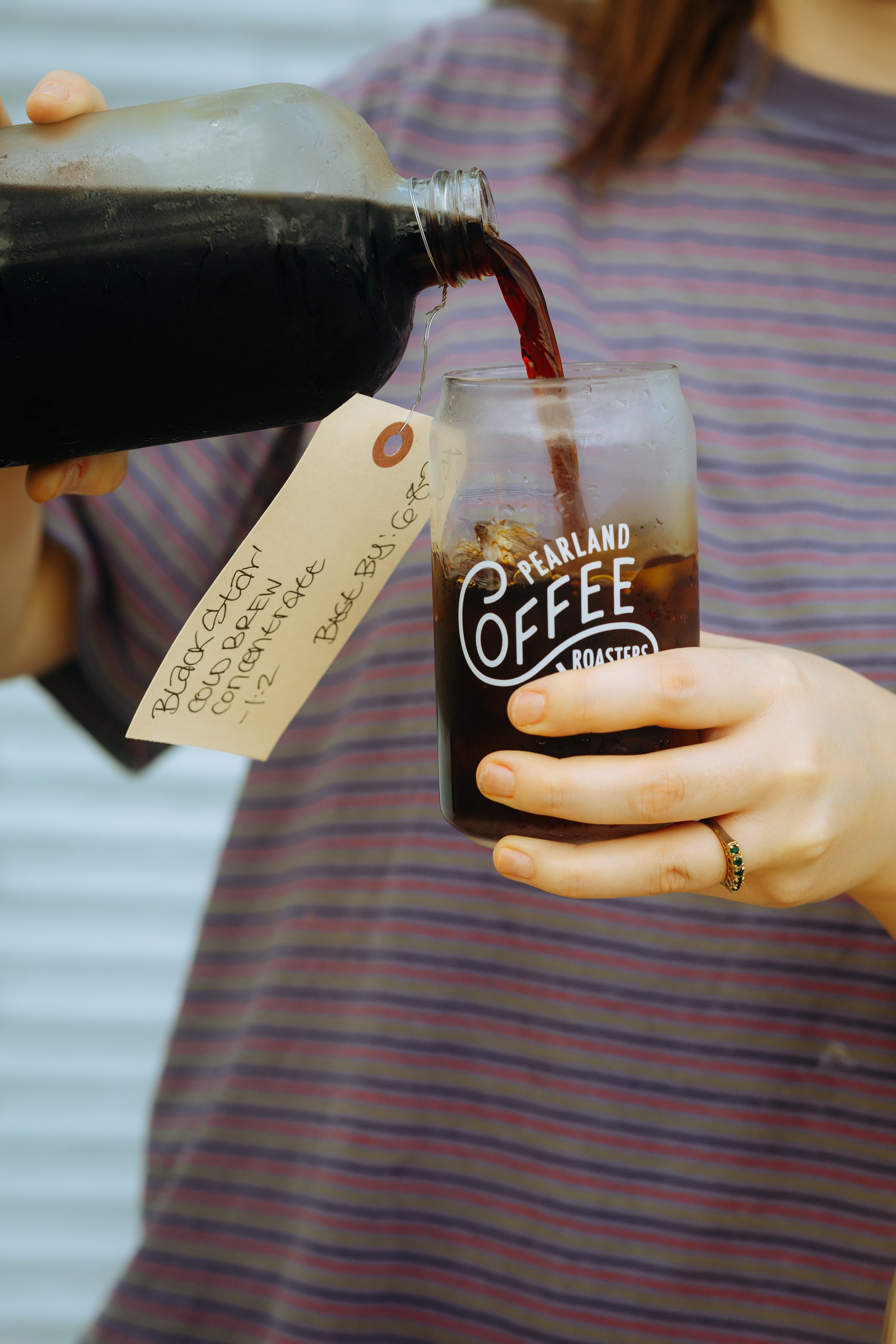 Person pouring cola from a bottle into a glass with ice, labeled 'Pearland Coffee Roasters'.