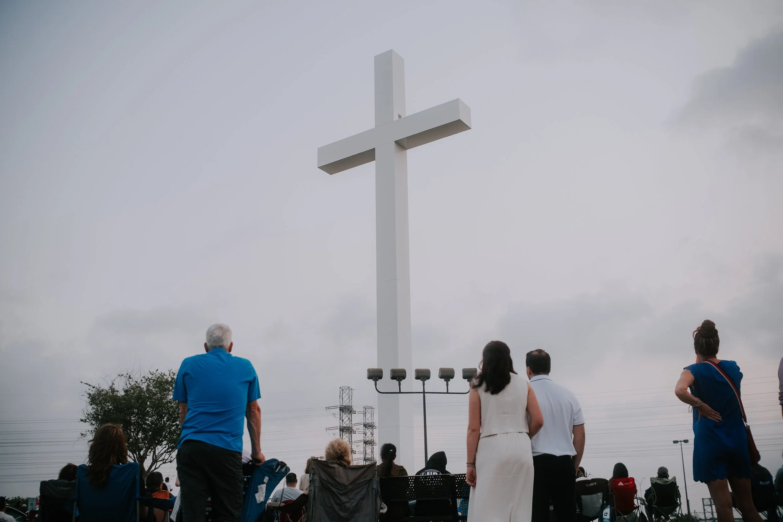 People gathered around an outdoor white cross monument, facing it with some seated and some standing, under a cloudy sky.
