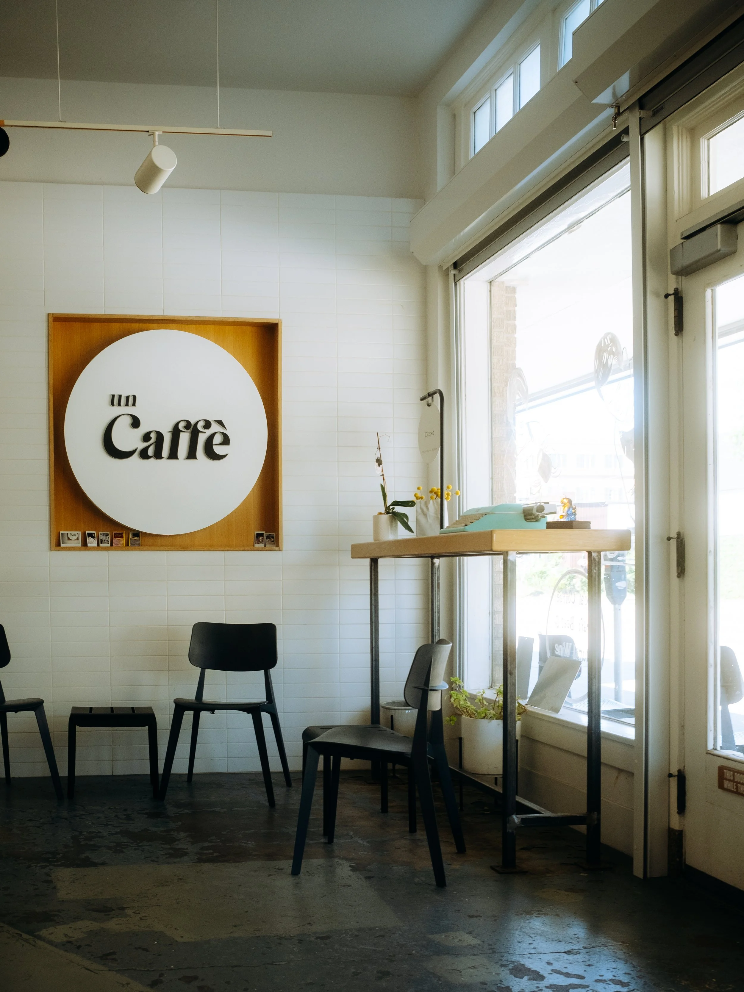 Interior of a coffee shop with a large round sign on the wall that says "un Caffè"; three black chairs, a small table with books and plants near large windows letting in sunlight.
