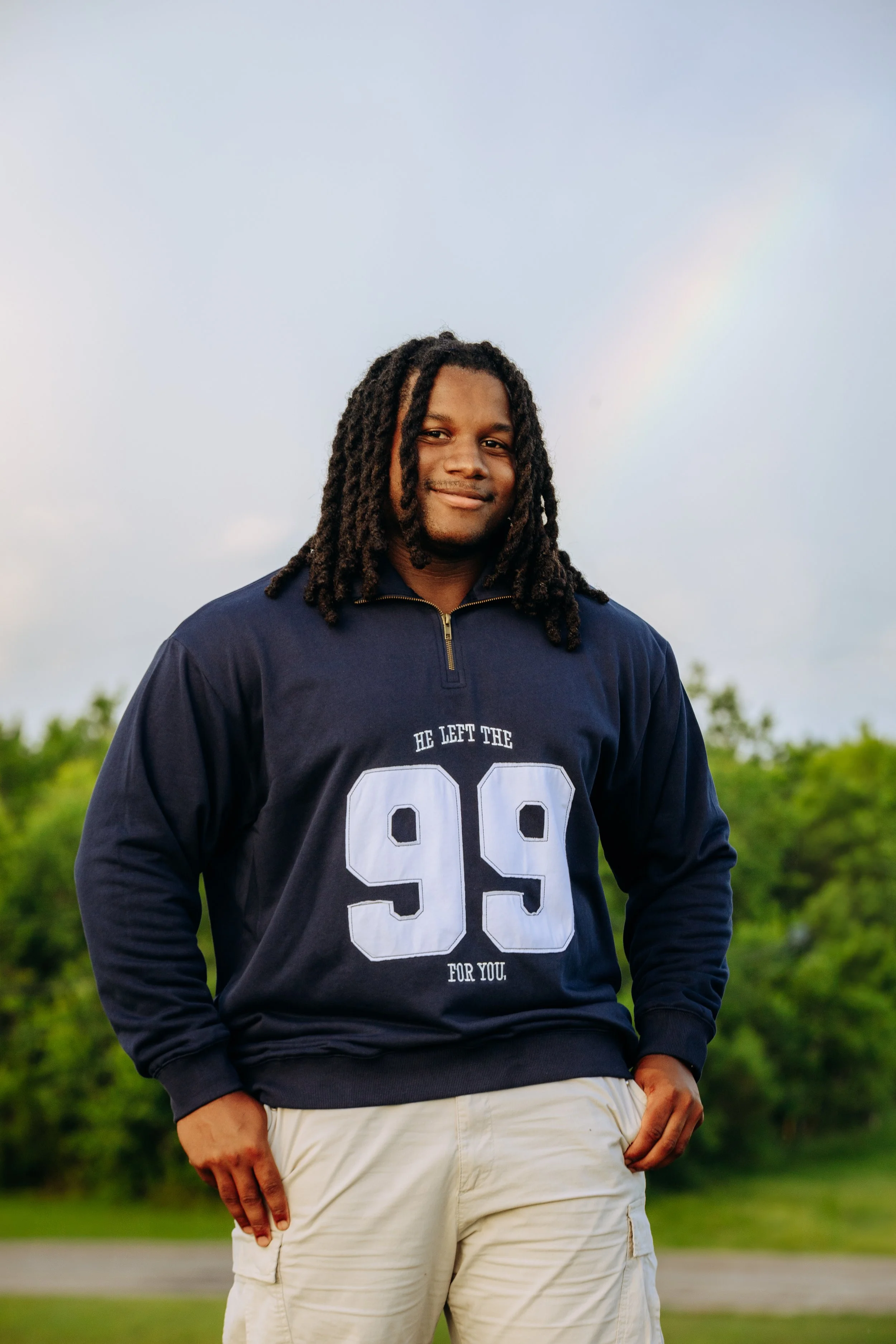 A young man with dreadlocks standing outdoors on a cloudy day with a faint rainbow in the background. He is wearing a navy blue sweatshirt with the number 99 and the words "He left the 99 for you" on it, and beige pants.