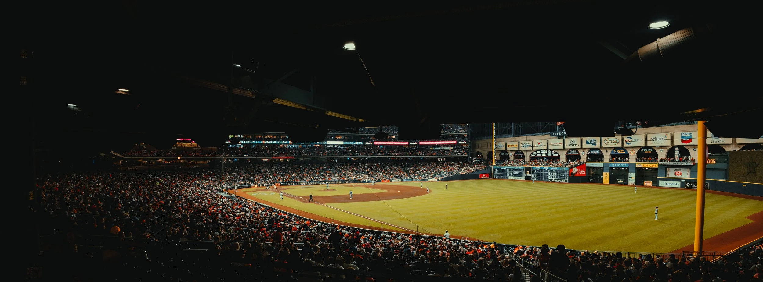 Interior of a baseball stadium filled with spectators watching a game, with players on the field and advertisements on the outfield walls